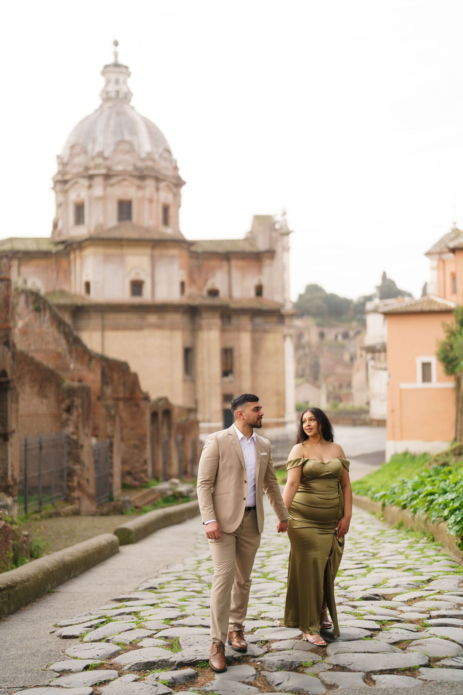 Couple walks on cobblestone path in Rome, woman in green gown, man in beige suit, historic church dome behind them.