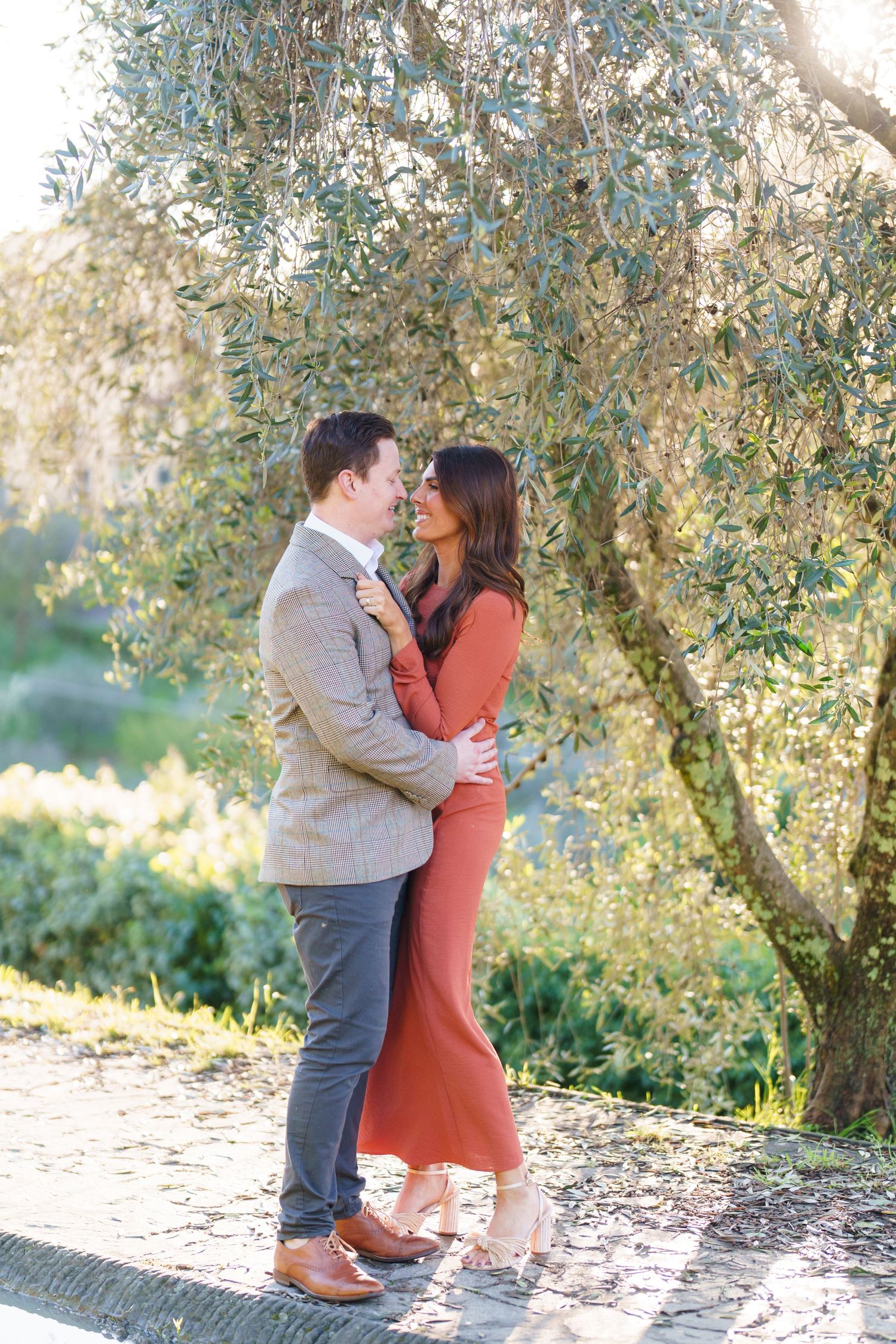 Couple embracing and gazing at each other under an olive tree, woman in rust dress, man in tweed blazer.
