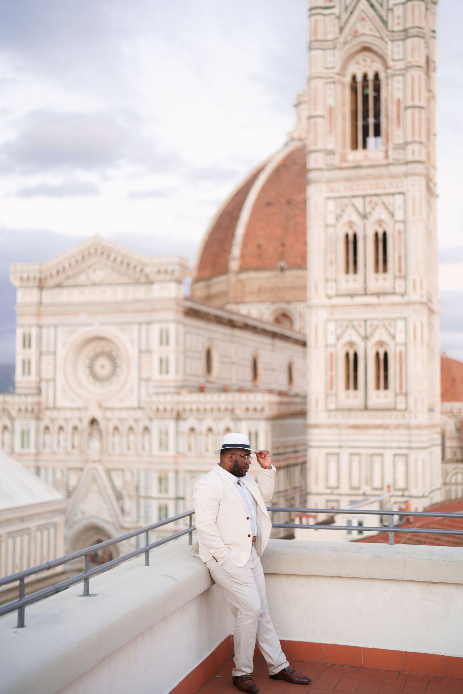 A well-dressed man in a white suit and hat poses on a rooftop terrace with Florence Cathedral in the background.