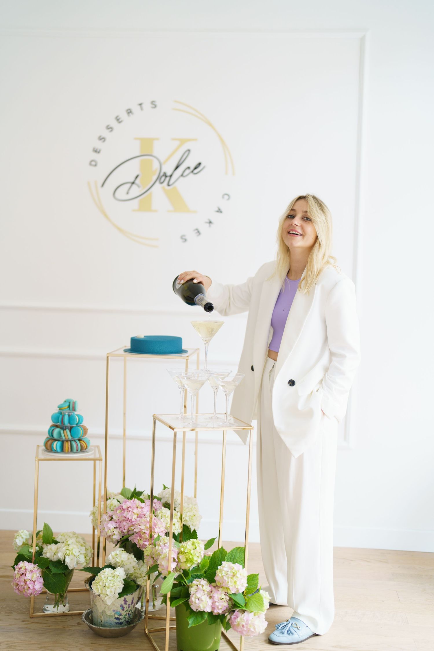 Blonde woman in white suit pouring champagne at K Dolce Desserts & Cakes bakery with macarons and flowers.
