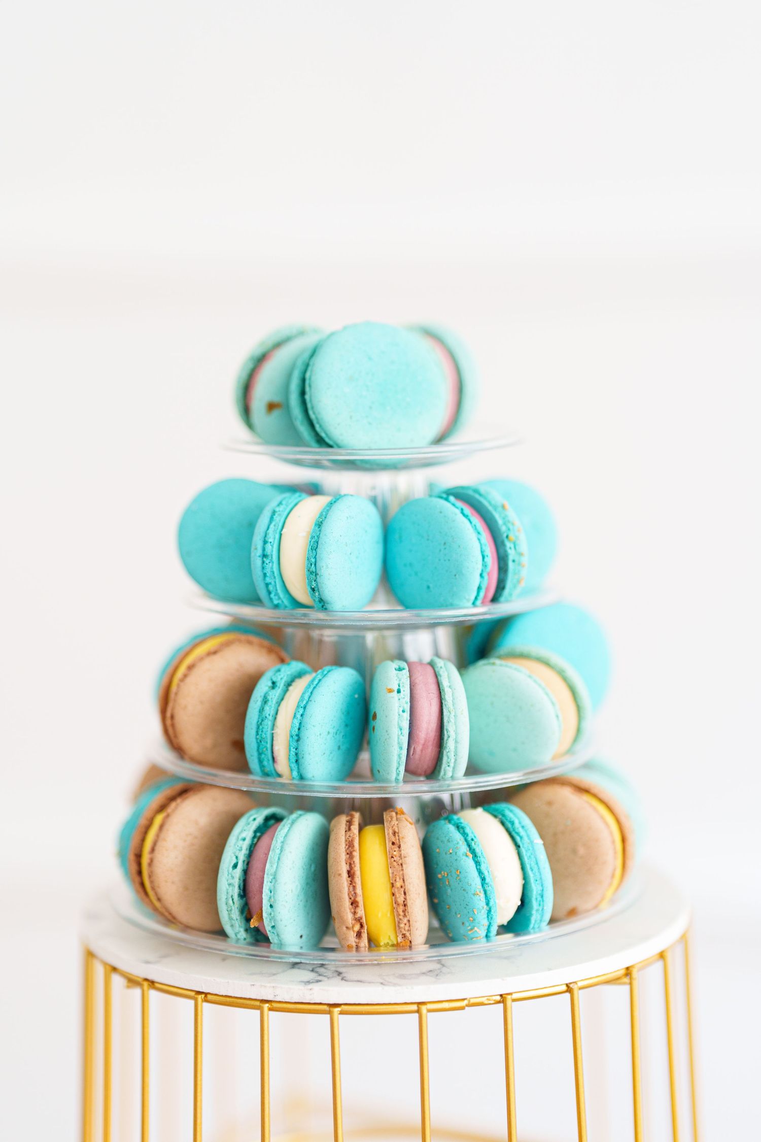 Elegant tiered display stand with colorful macarons in turquoise, beige, and yellow on a gold base.