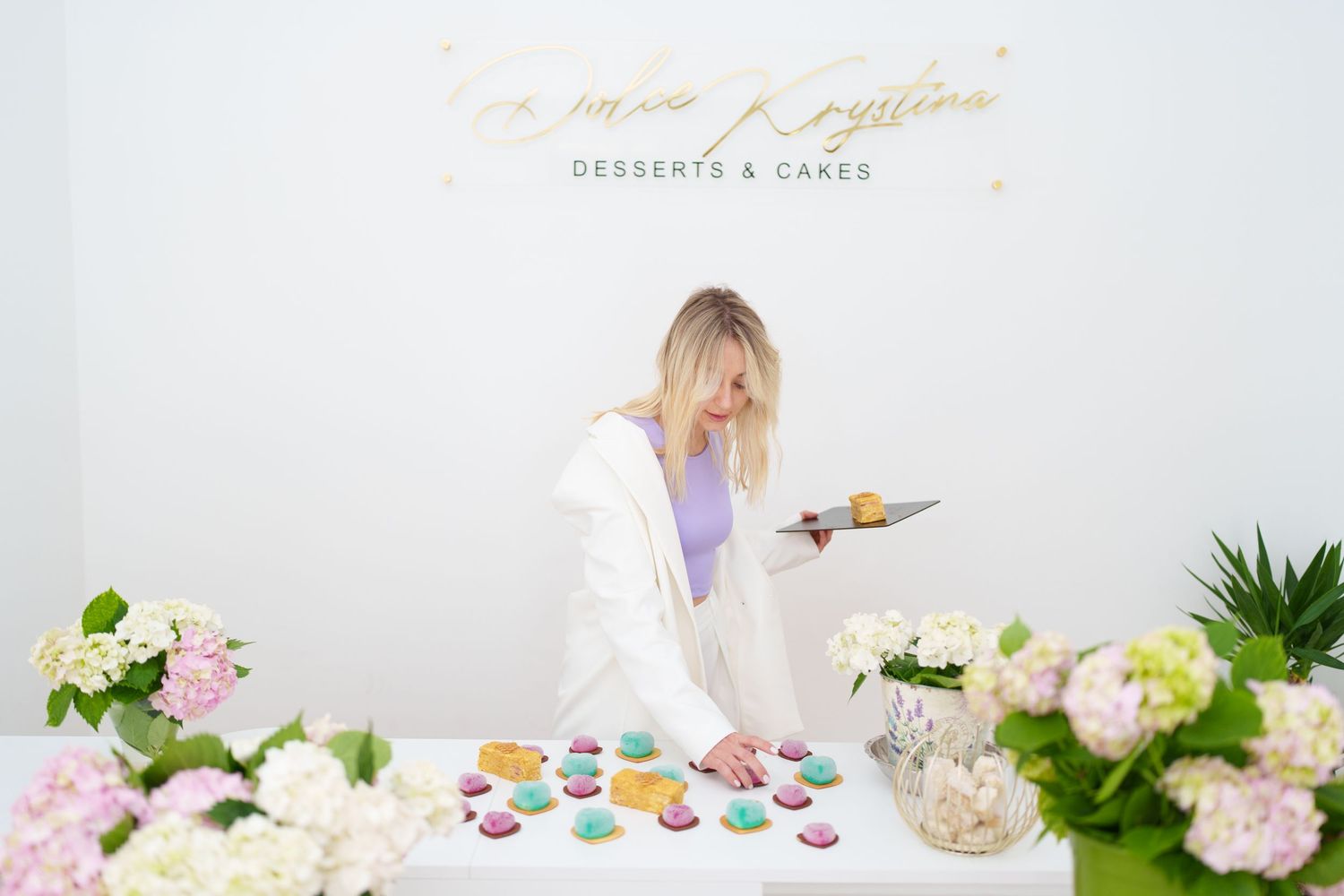 Woman arranging colorful macarons on a white table with floral decor at a desserts and cakes shop.