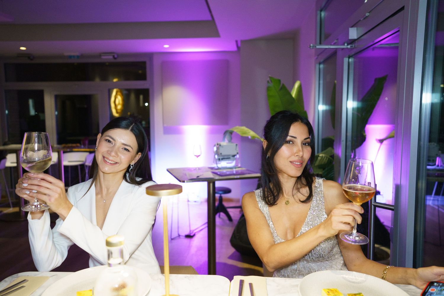 Two women toasting with wine glasses at an elegant restaurant with purple ambient lighting.