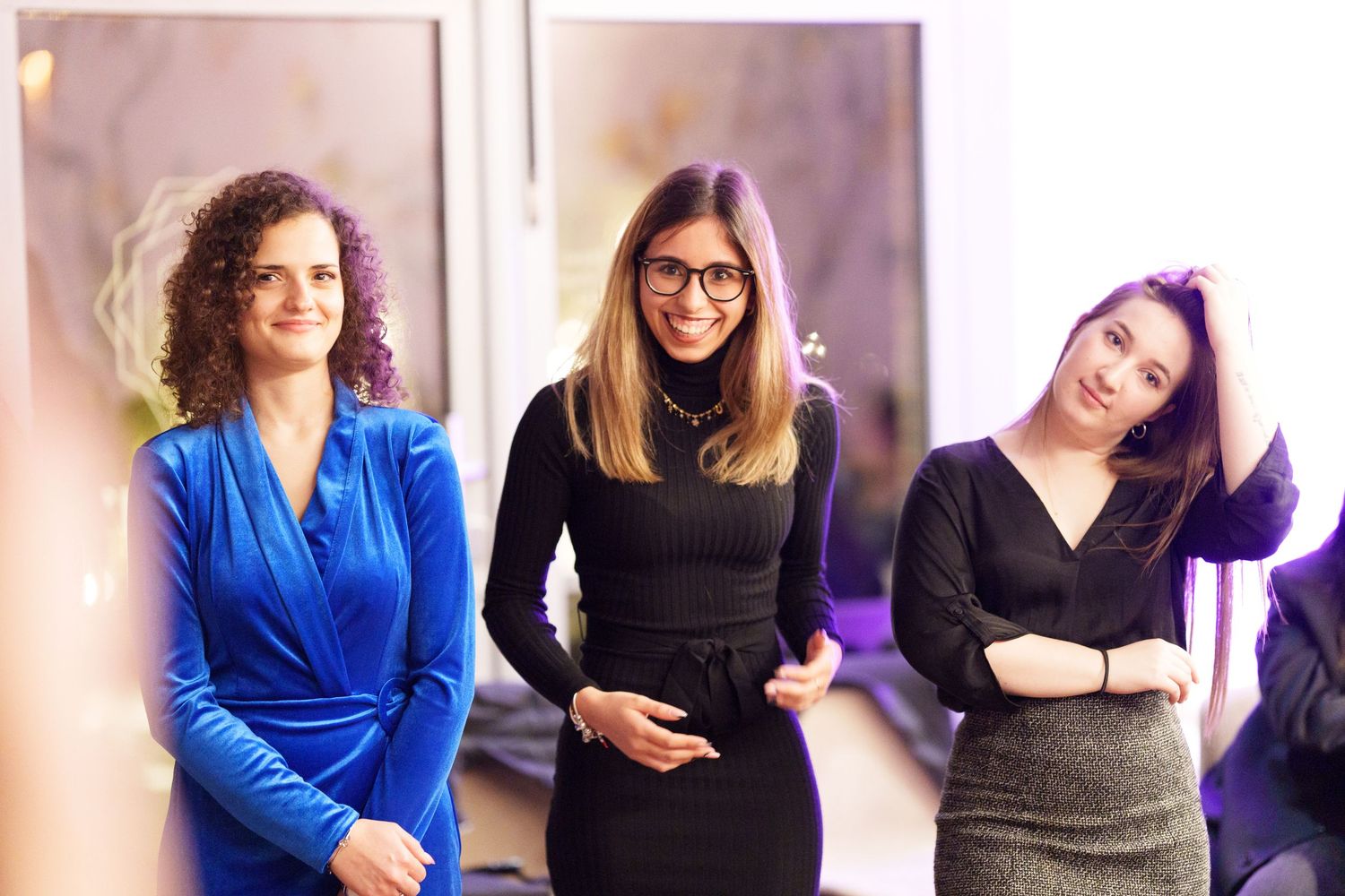 Three professional women smiling and posing together indoors, dressed in elegant blue, black, and dark outfits.