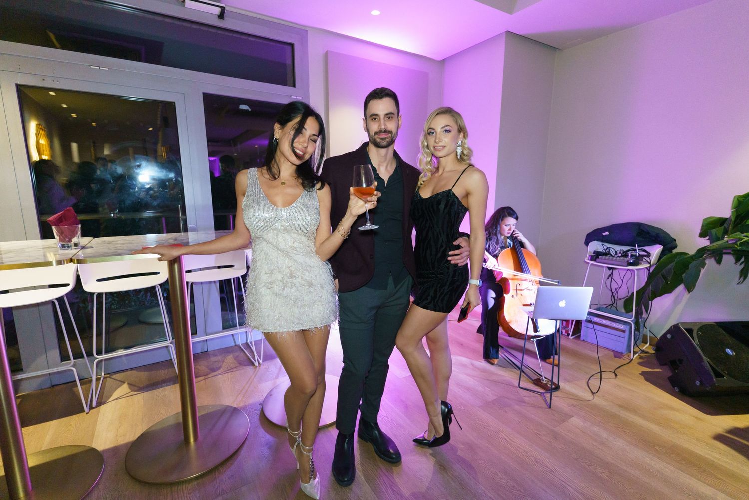 Two women and a man pose with drinks at an indoor party, with a DJ visible in the background.