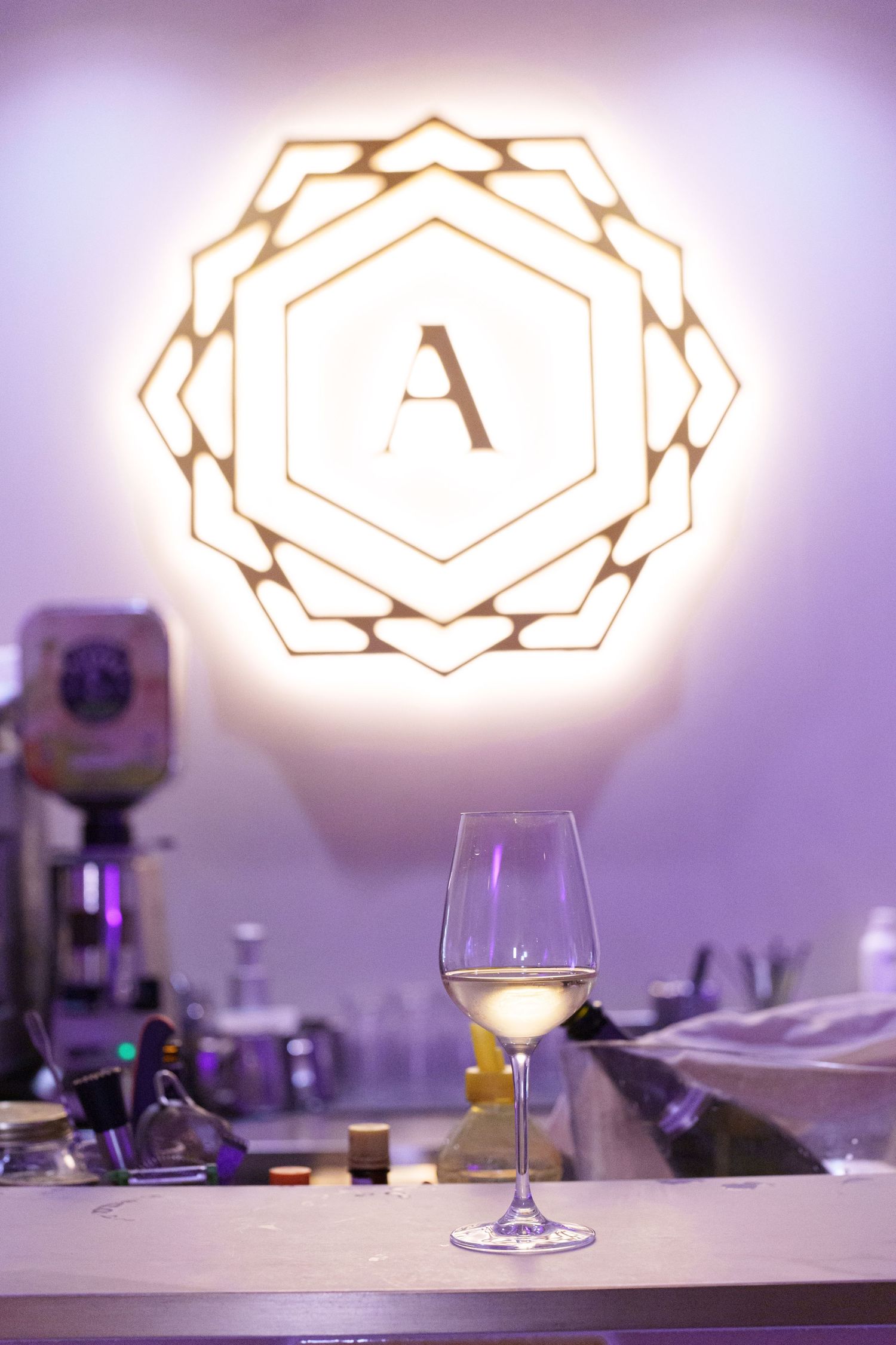A wine glass with white wine sits on a bar counter beneath an illuminated hexagonal logo sign with letter A.