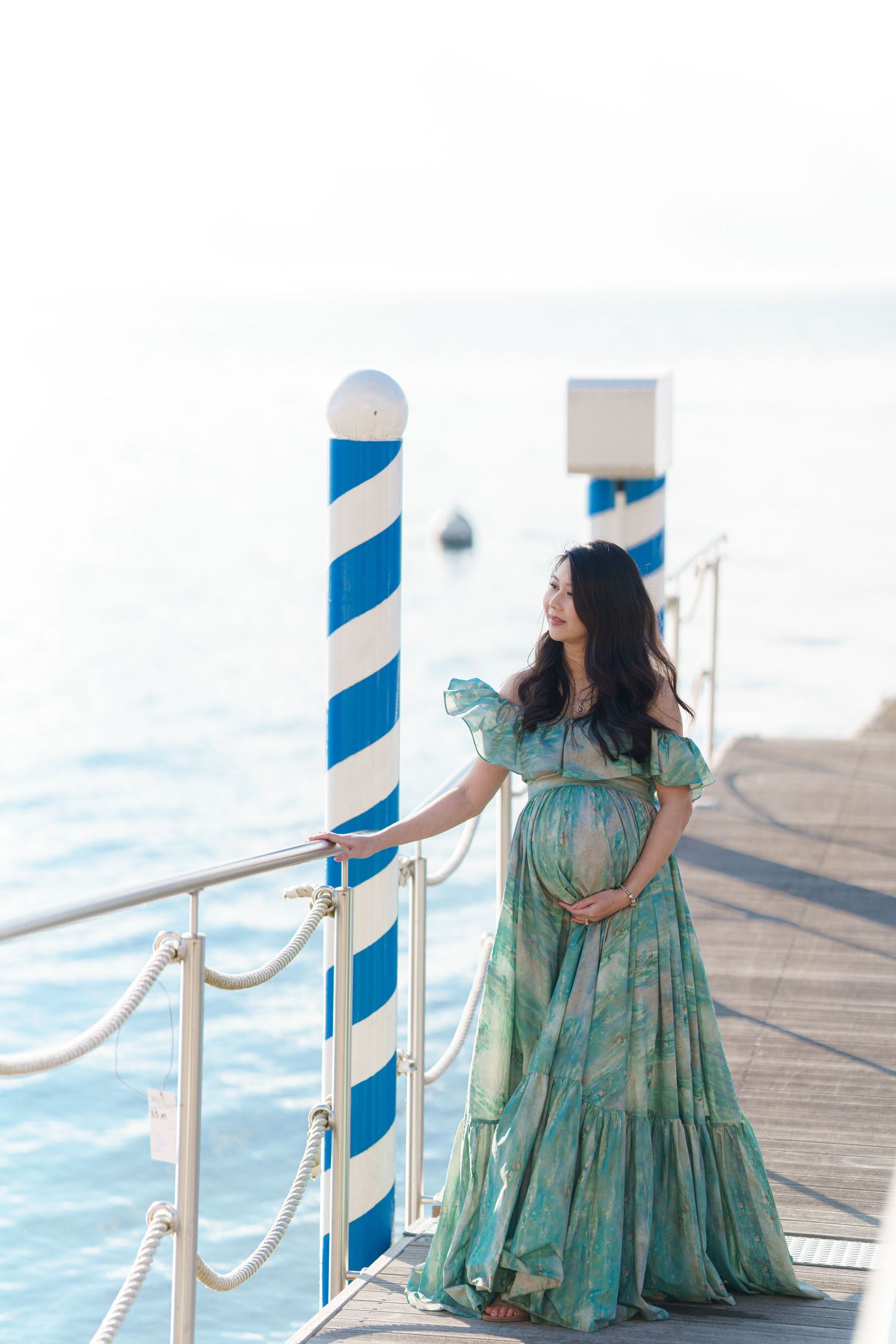 Pregnant woman in green floral dress standing on a pier by the sea, cradling her baby bump near blue striped poles.