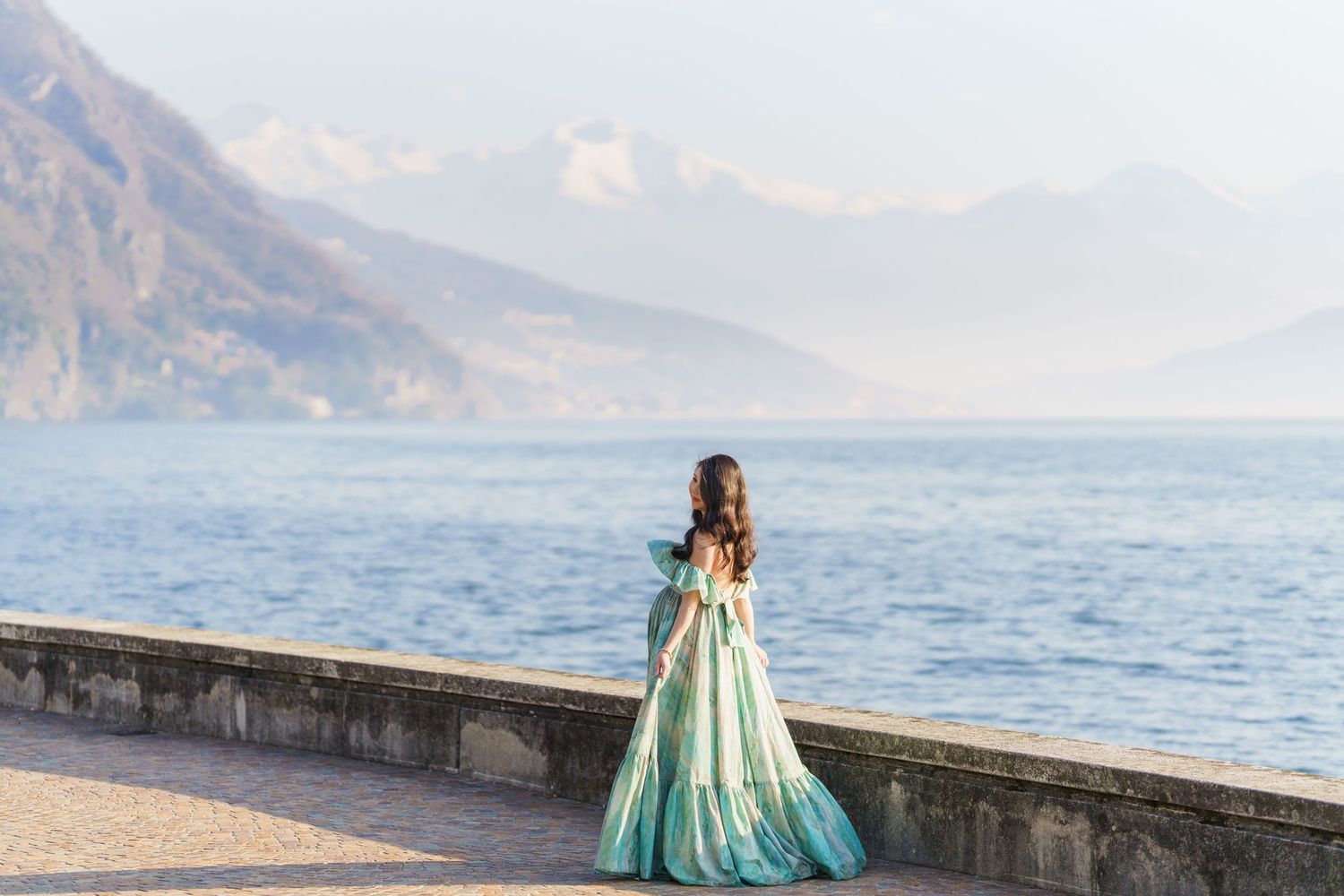 A woman in a flowing teal ball gown stands by a lakeside promenade with misty mountains in the background.