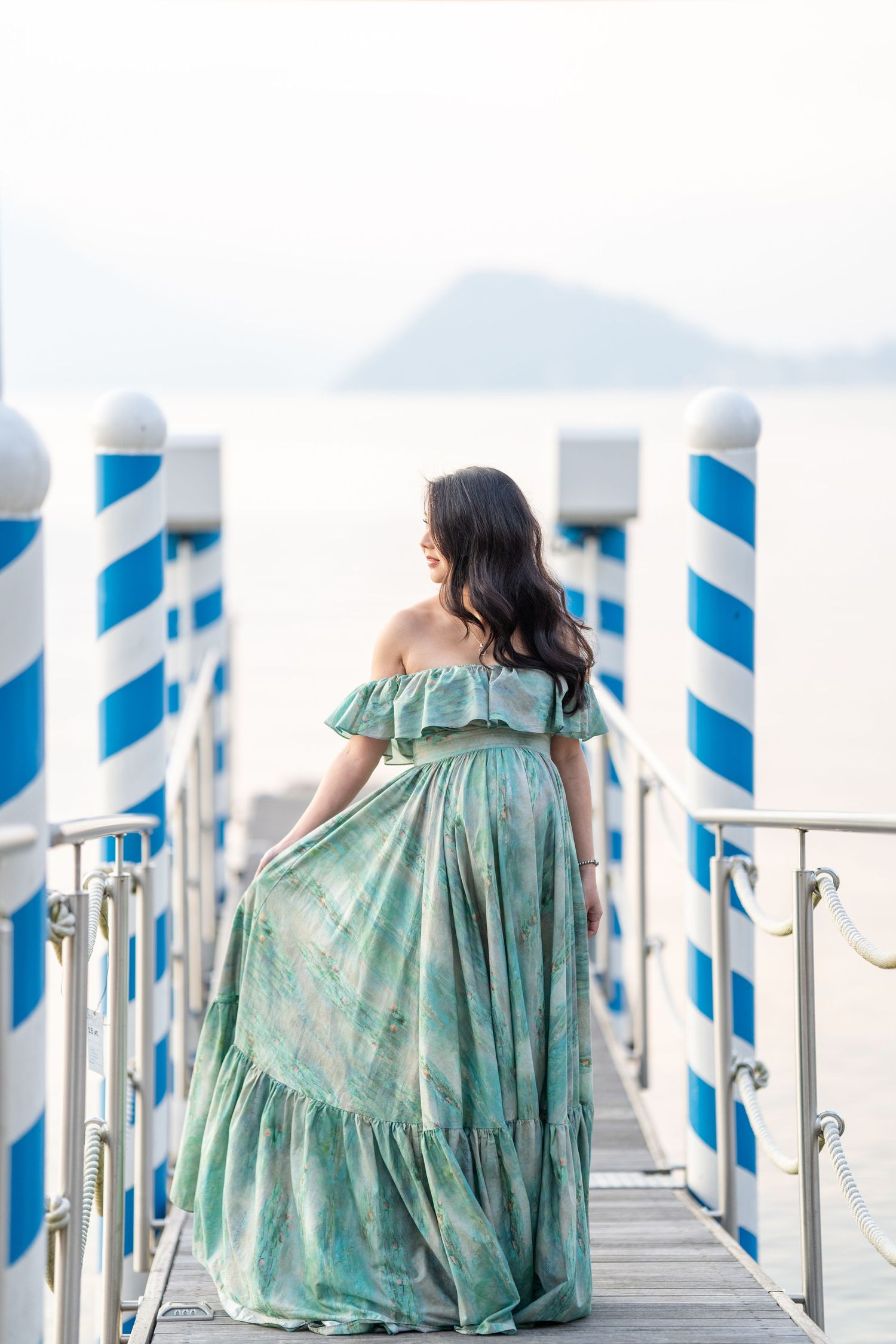 Woman in green off-shoulder maxi dress standing on a blue and white pier overlooking calm ocean waters.