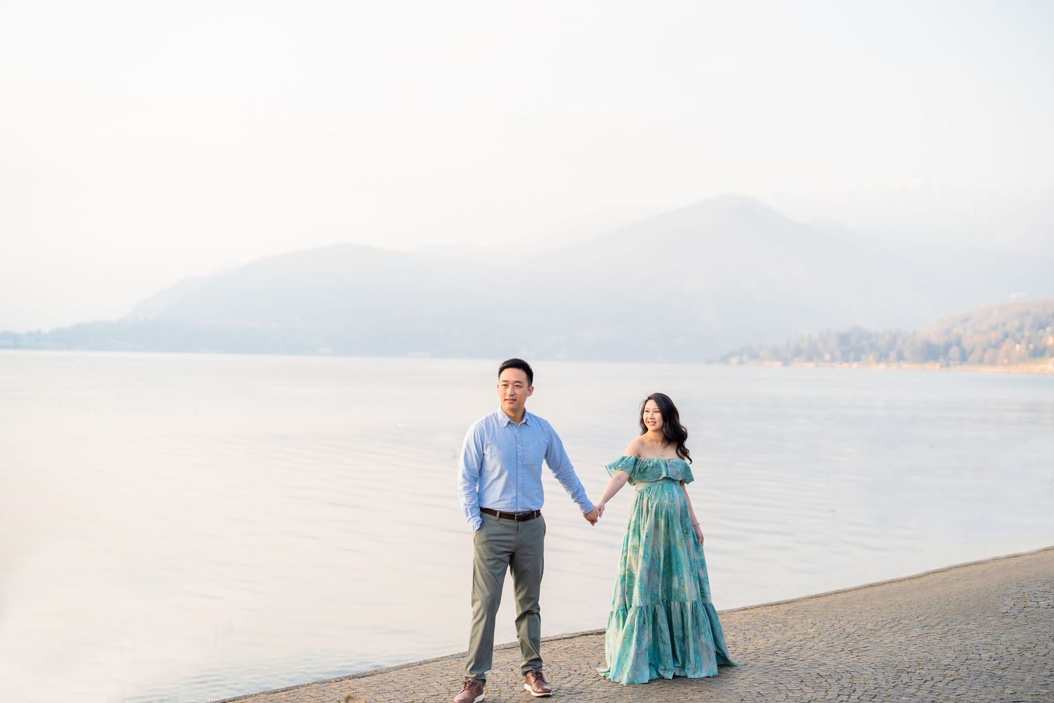 Couple holding hands by a serene lakeside with misty mountains, woman in teal gown, man in blue shirt.