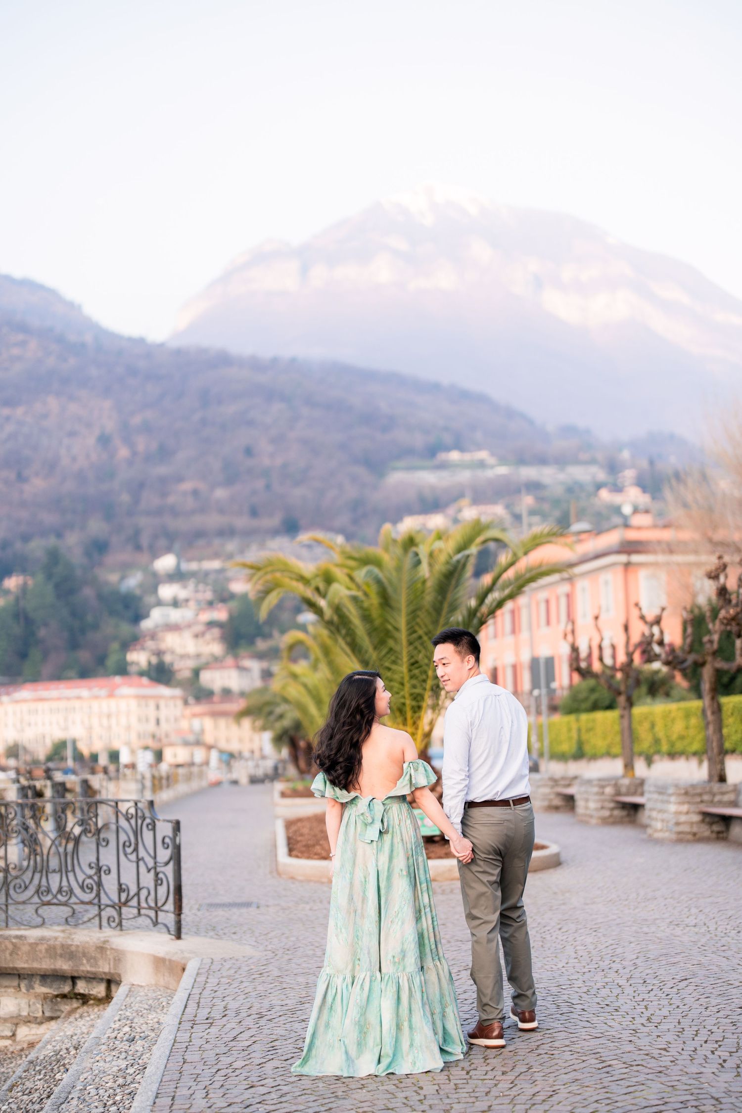 Couple holding hands on cobblestone promenade with mountain backdrop and palm trees at Lake Como, Italy.