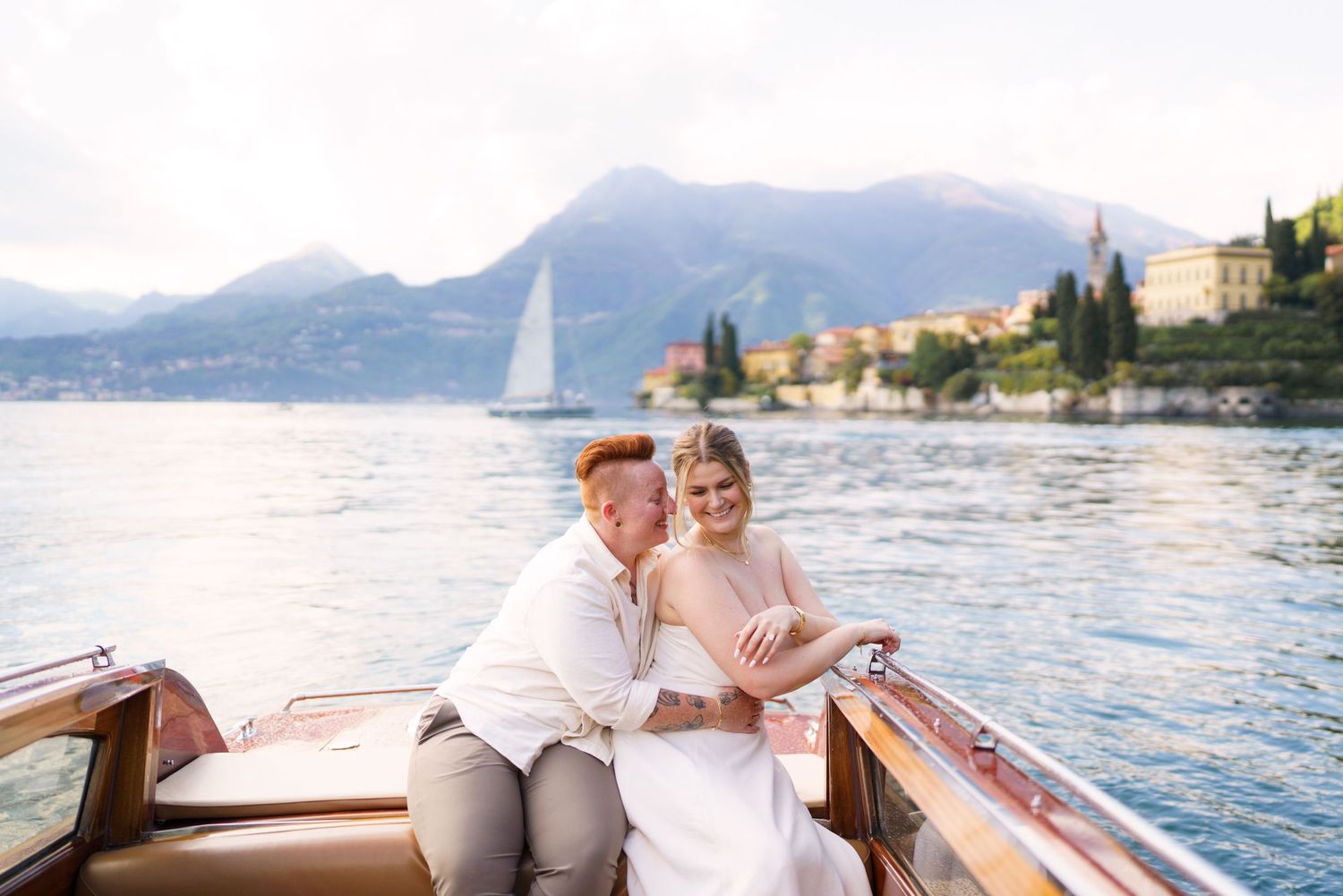 Couple embracing on a classic wooden boat on Lake Como, Italy, with mountains and a sailboat in the background.