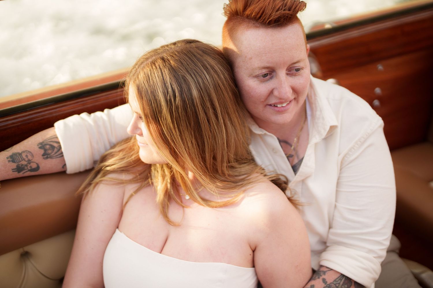 Two brides share a tender moment on a boat, one with red hair and tattoos smiling warmly at the other.