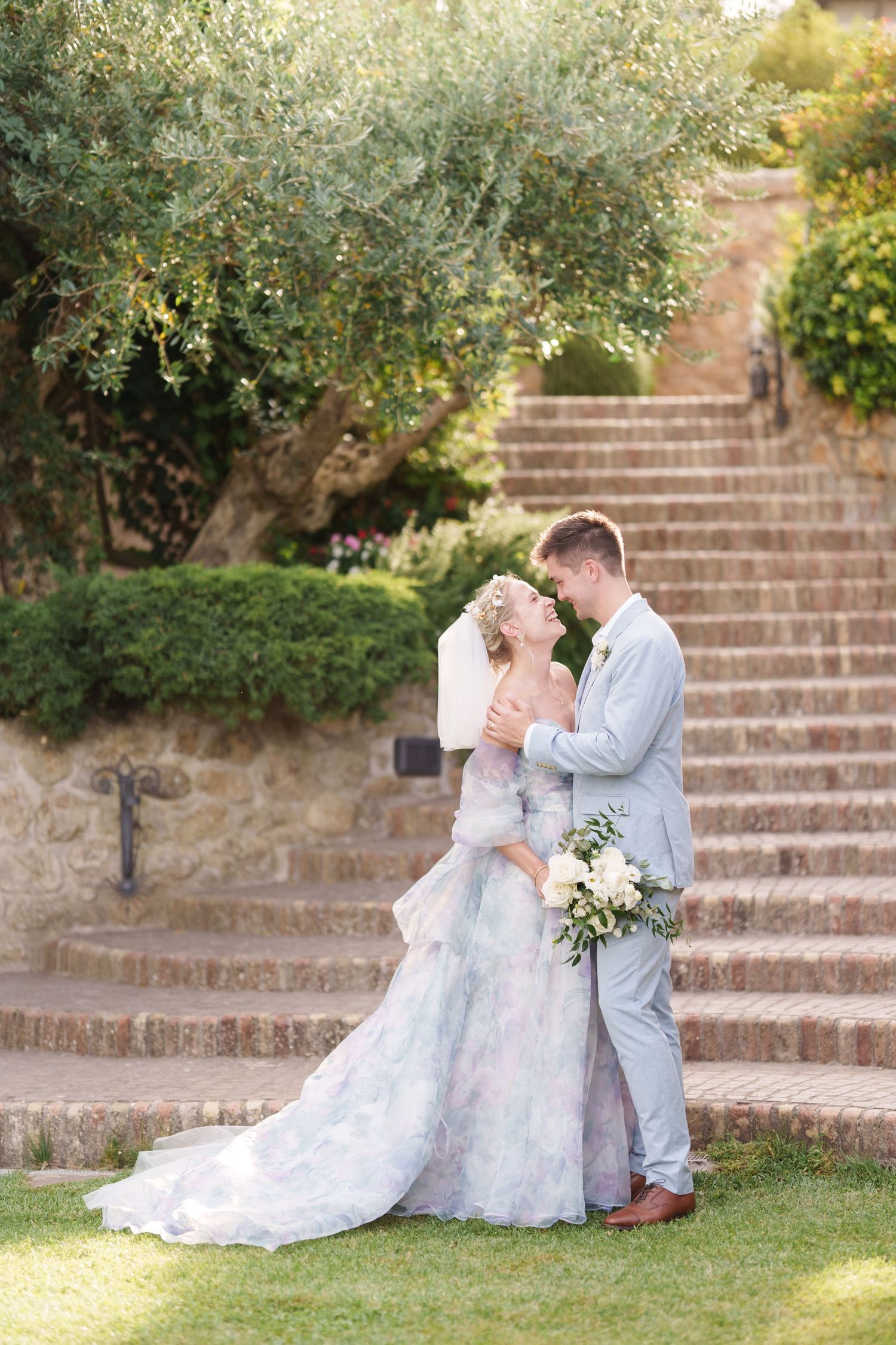 Bride in floral gown and groom in gray suit embrace near stone steps surrounded by lush greenery.