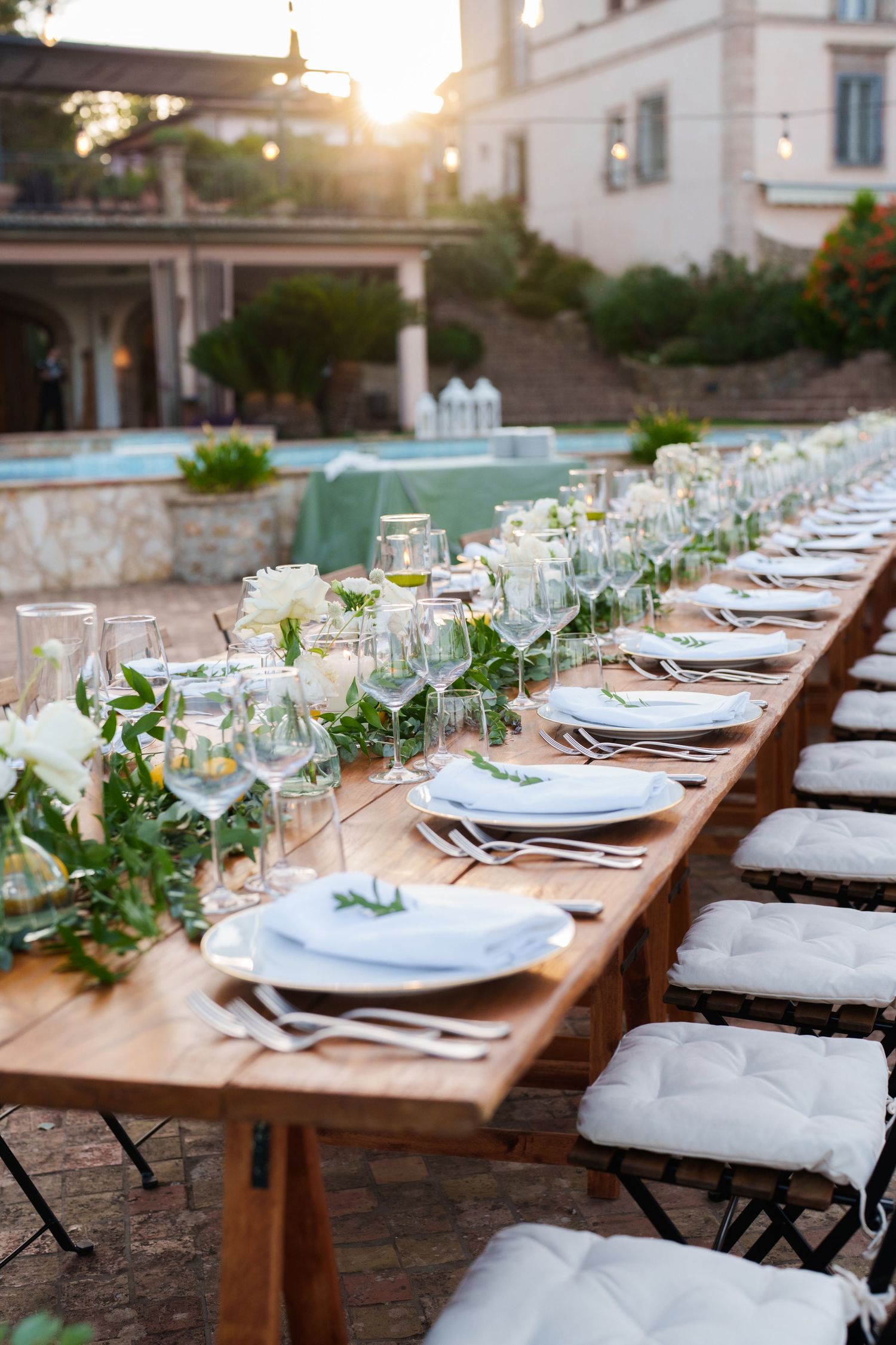 Elegant outdoor wedding reception table set with white flowers, greenery, glassware, and white cushioned chairs at golden hour.