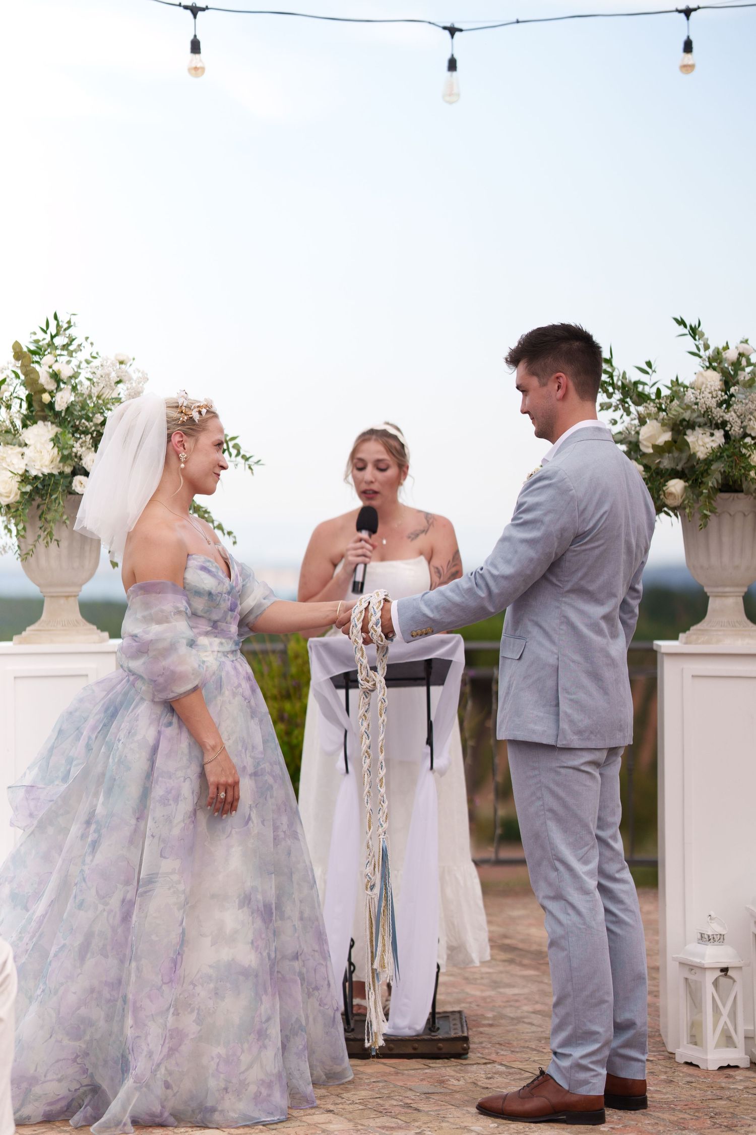 Bride and groom exchange vows outdoors with officiant, floral arrangements, and string lights at elegant wedding ceremony.