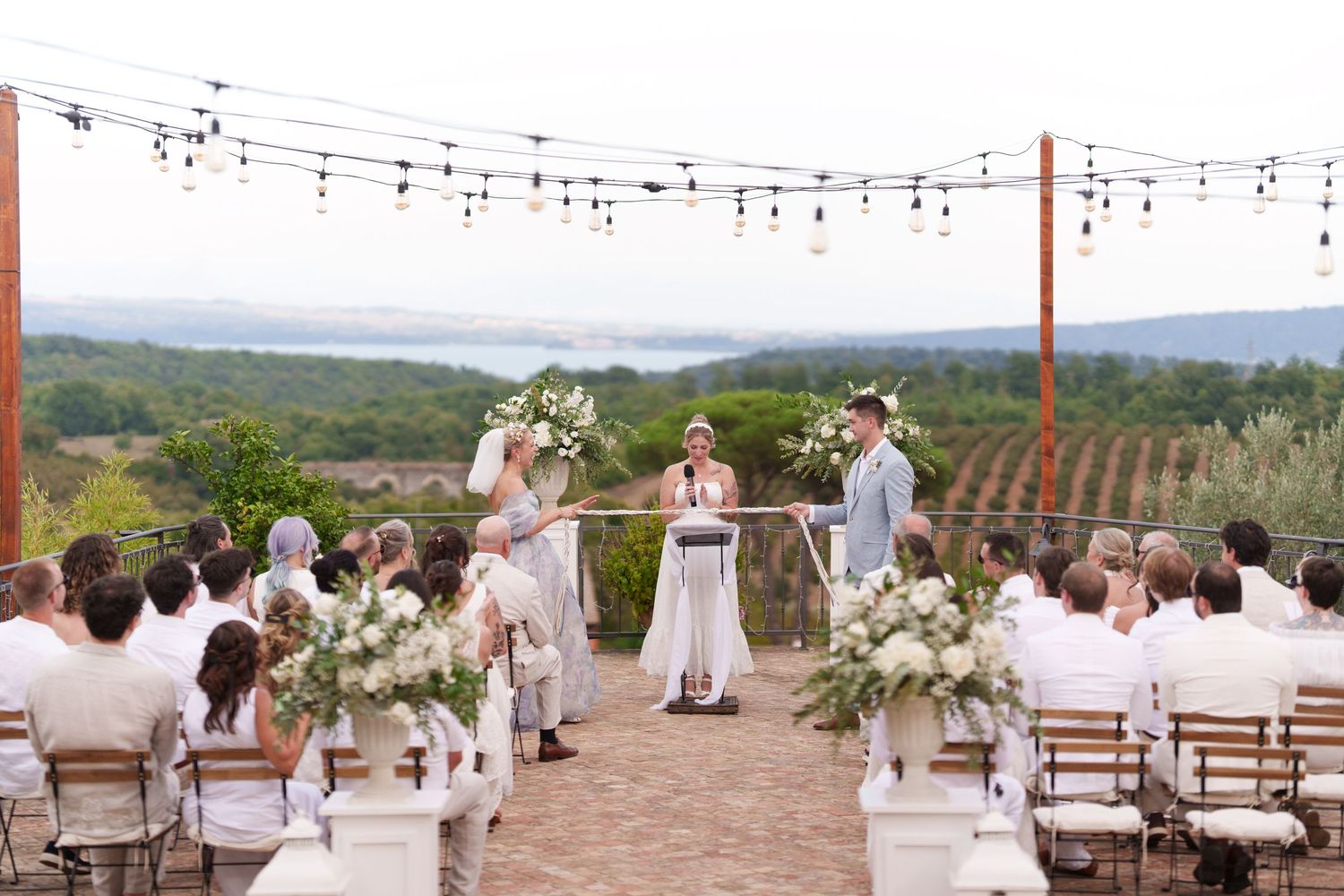 Outdoor vineyard wedding ceremony with string lights, white floral arrangements, and scenic mountain lake views.