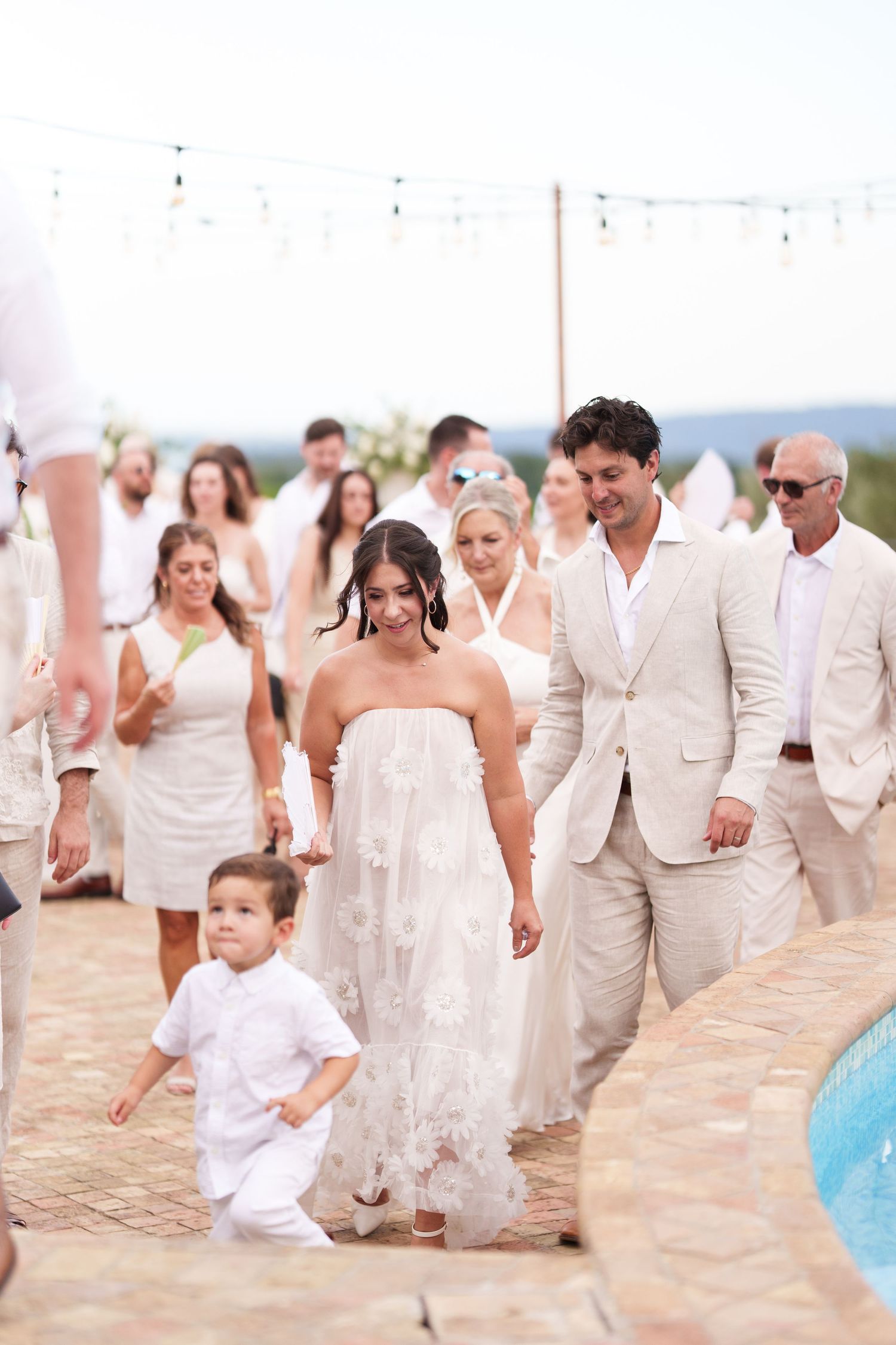 Bride and groom walk with young boy at outdoor wedding, guests in white attire near pool.