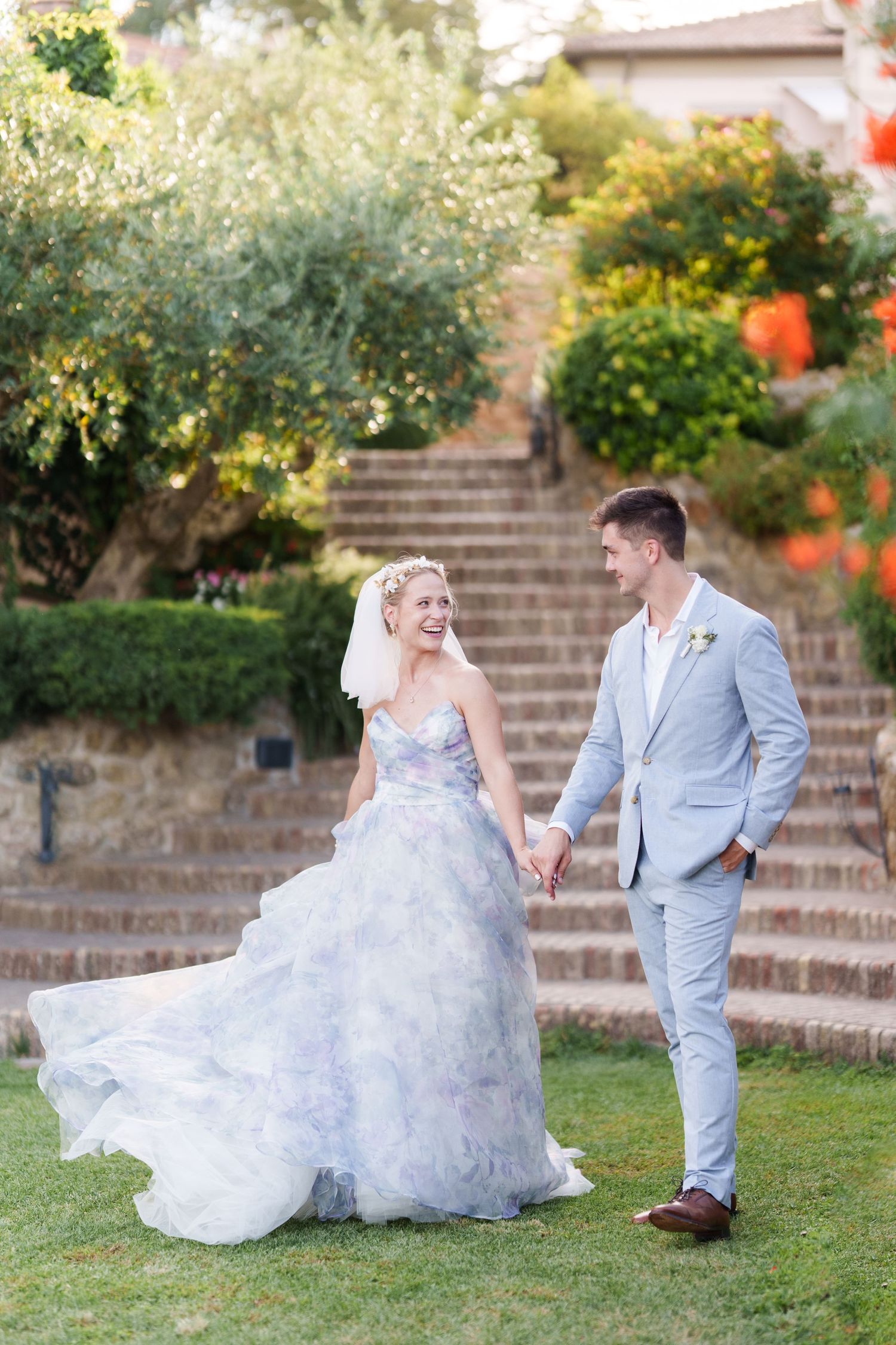 Bride in floral gown and groom in light blue suit walk hand in hand laughing in lush garden with stone steps.