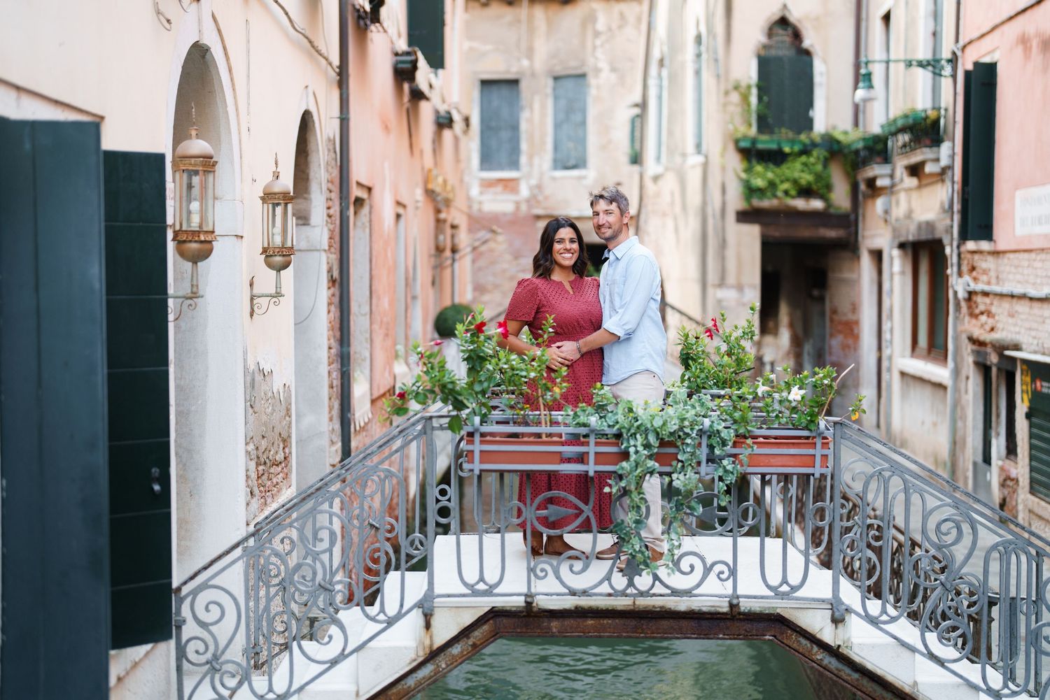 Couple smiling on ornate bridge over Venetian canal surrounded by lush potted plants in Venice, Italy.