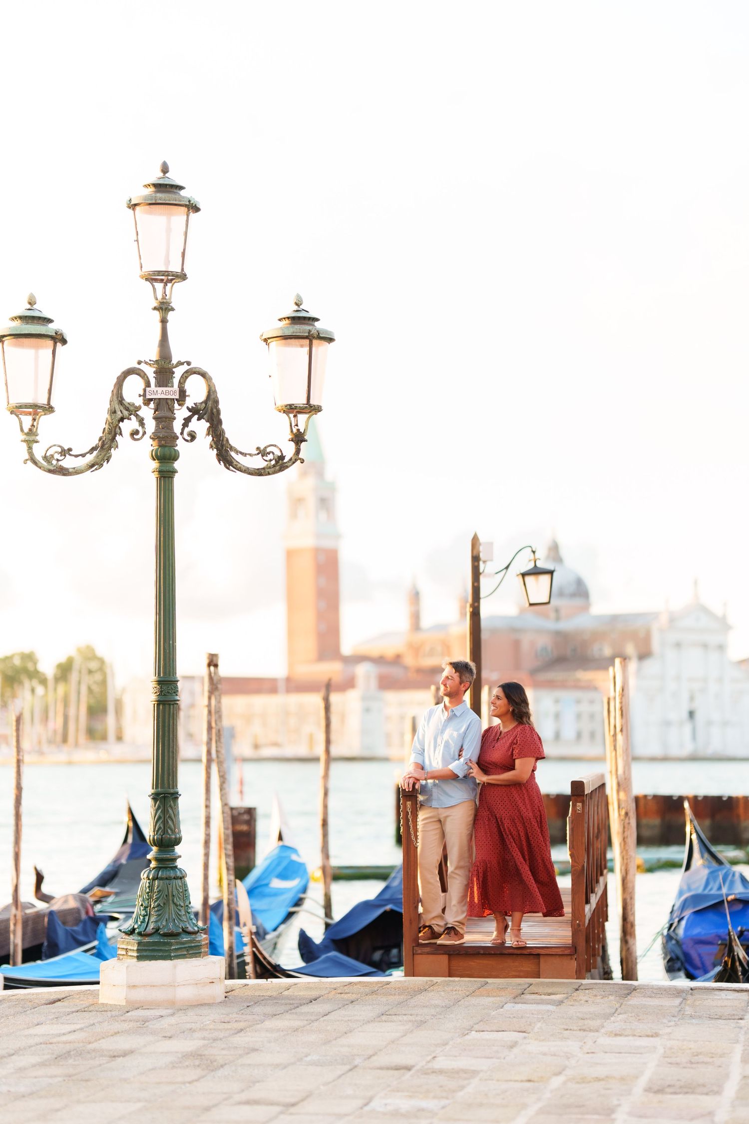 Couple embracing on Venice waterfront with gondolas and San Giorgio Maggiore church in golden morning light.