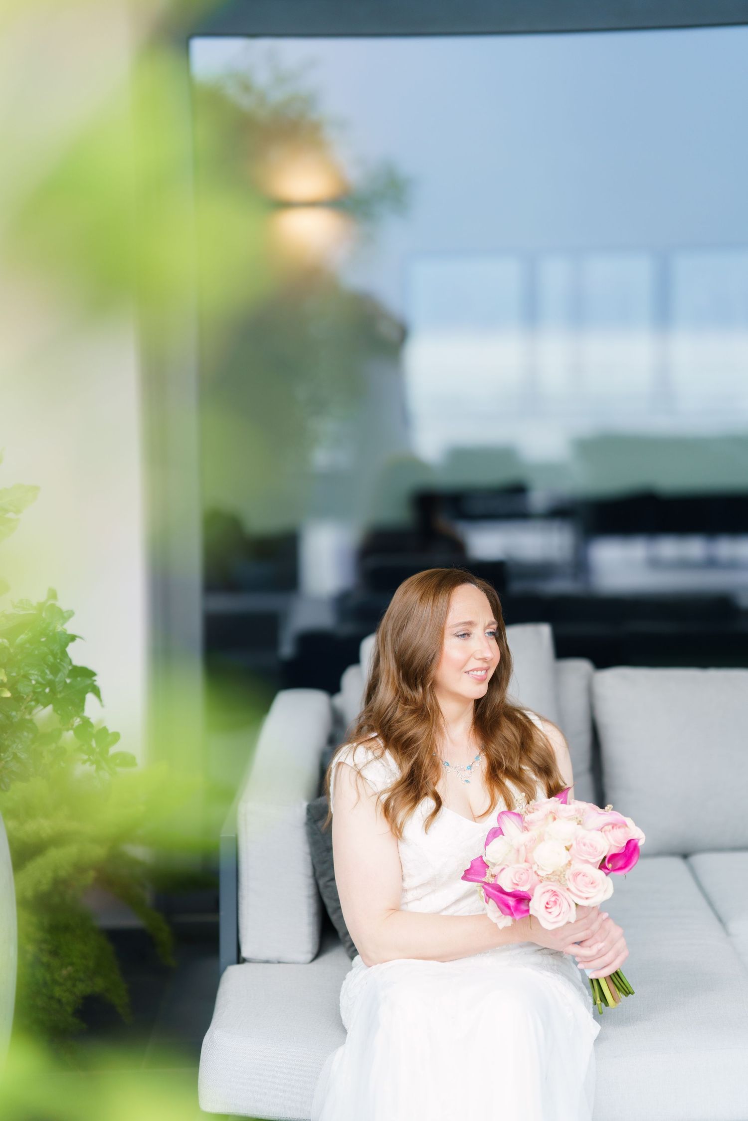 Bride in white dress sitting on outdoor sofa holding pink rose bouquet, smiling softly in elegant wedding setting.
