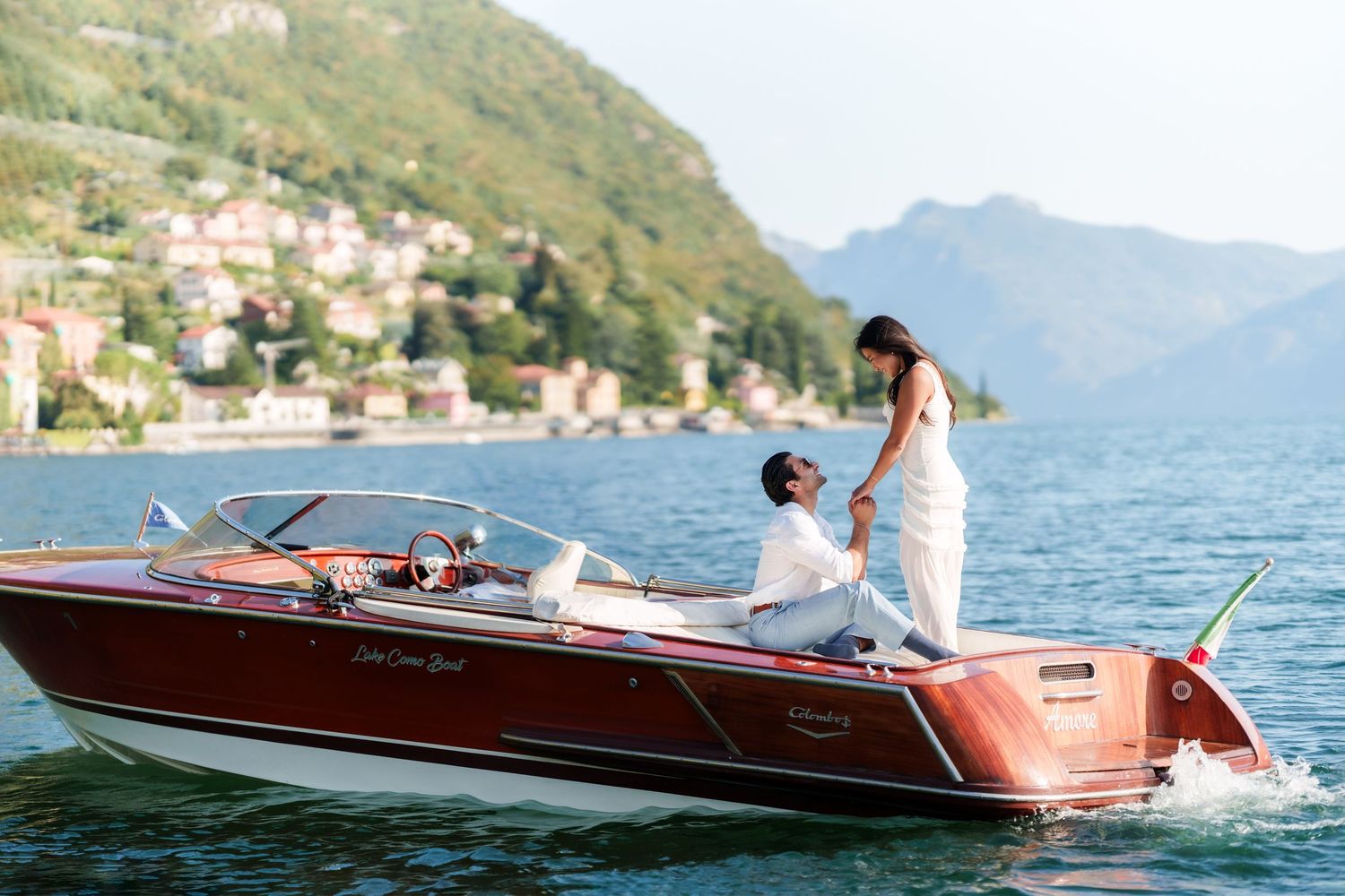 Couple on a classic wooden speedboat on Lake Como, Italy, with mountains and village in the background.
