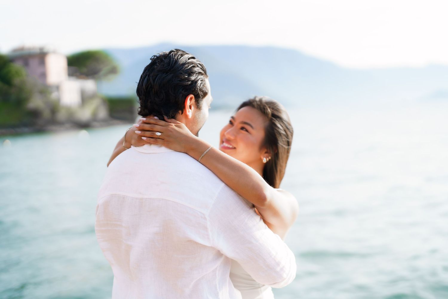 Couple embracing and smiling by a scenic lake with mountains and a village in the background.