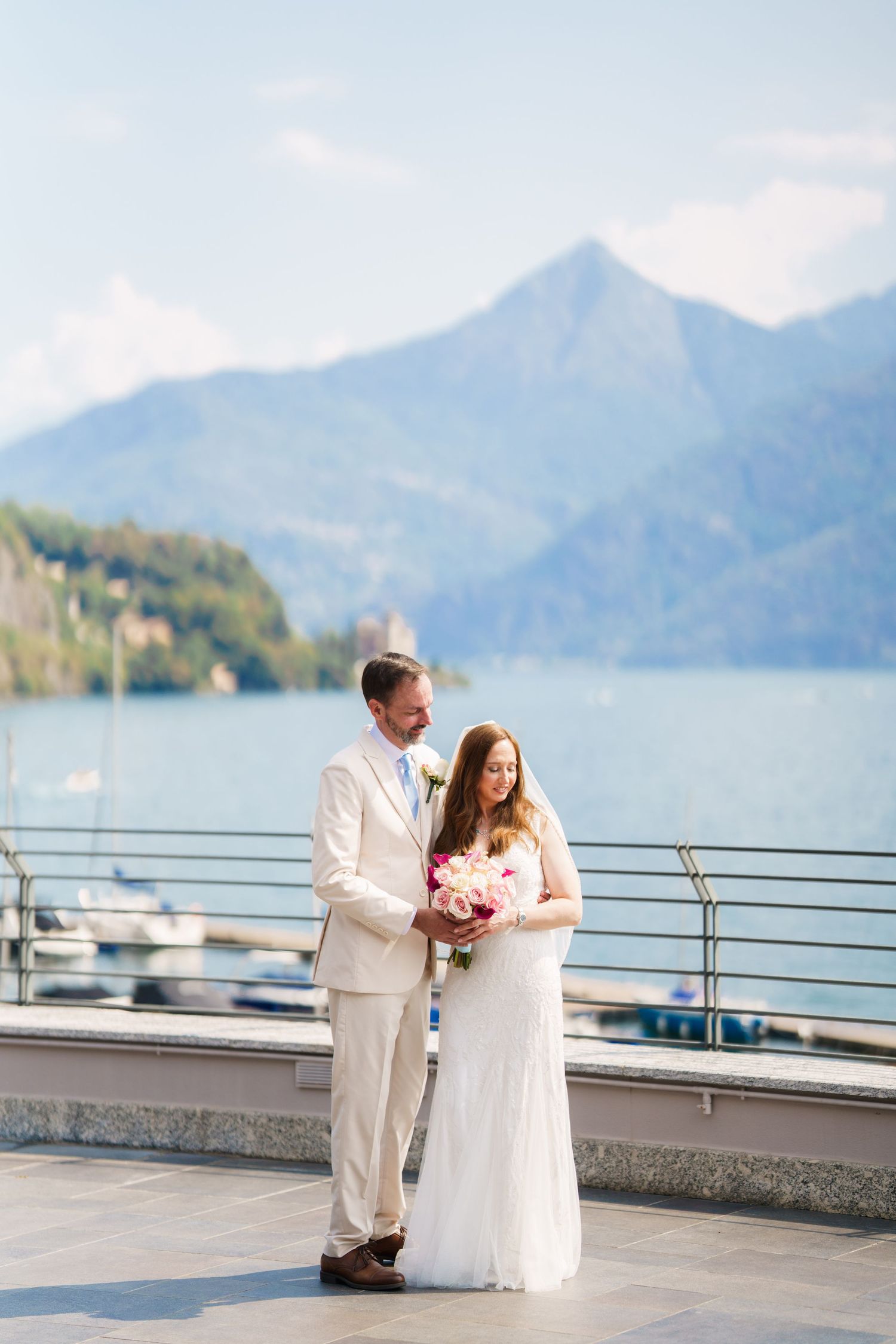 Bride and groom pose on lakeside terrace with mountain backdrop, bride holding pink bouquet in white dress.