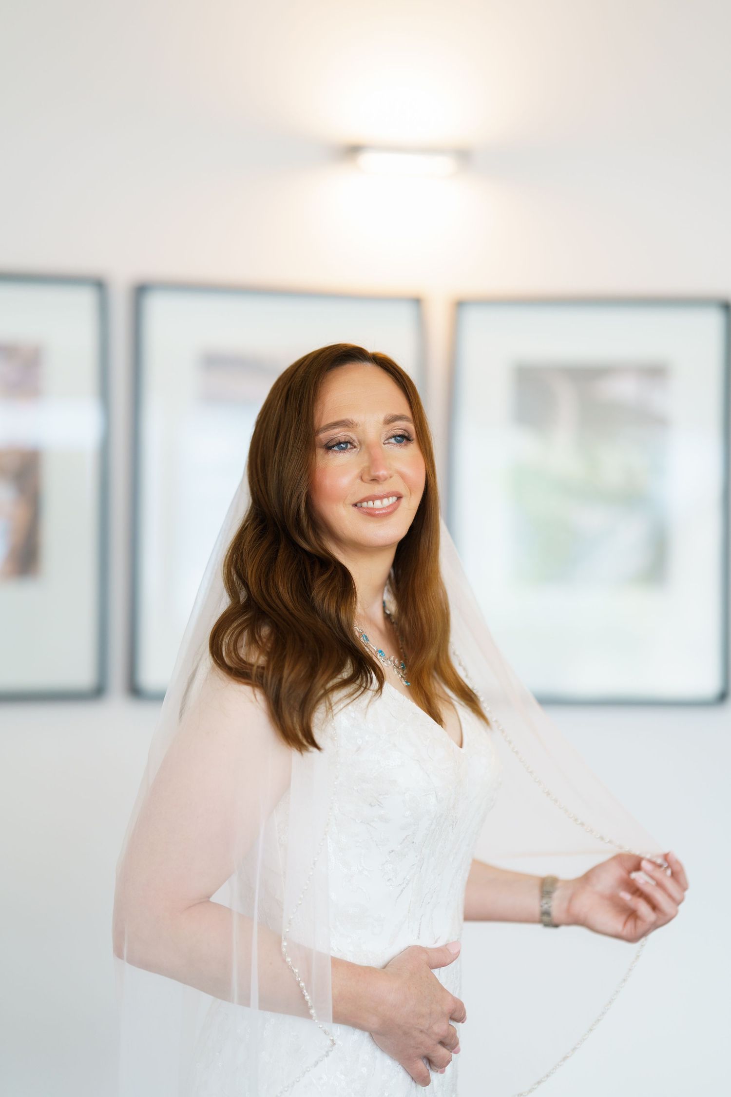 Smiling bride in white lace gown and veil holding dress, brown hair, freckles, standing indoors near framed photos.