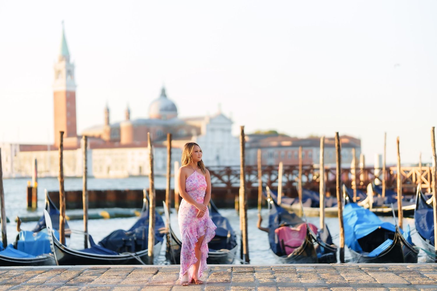 Woman in pink dress stands by gondolas on Venice waterfront with San Giorgio Maggiore island in background.