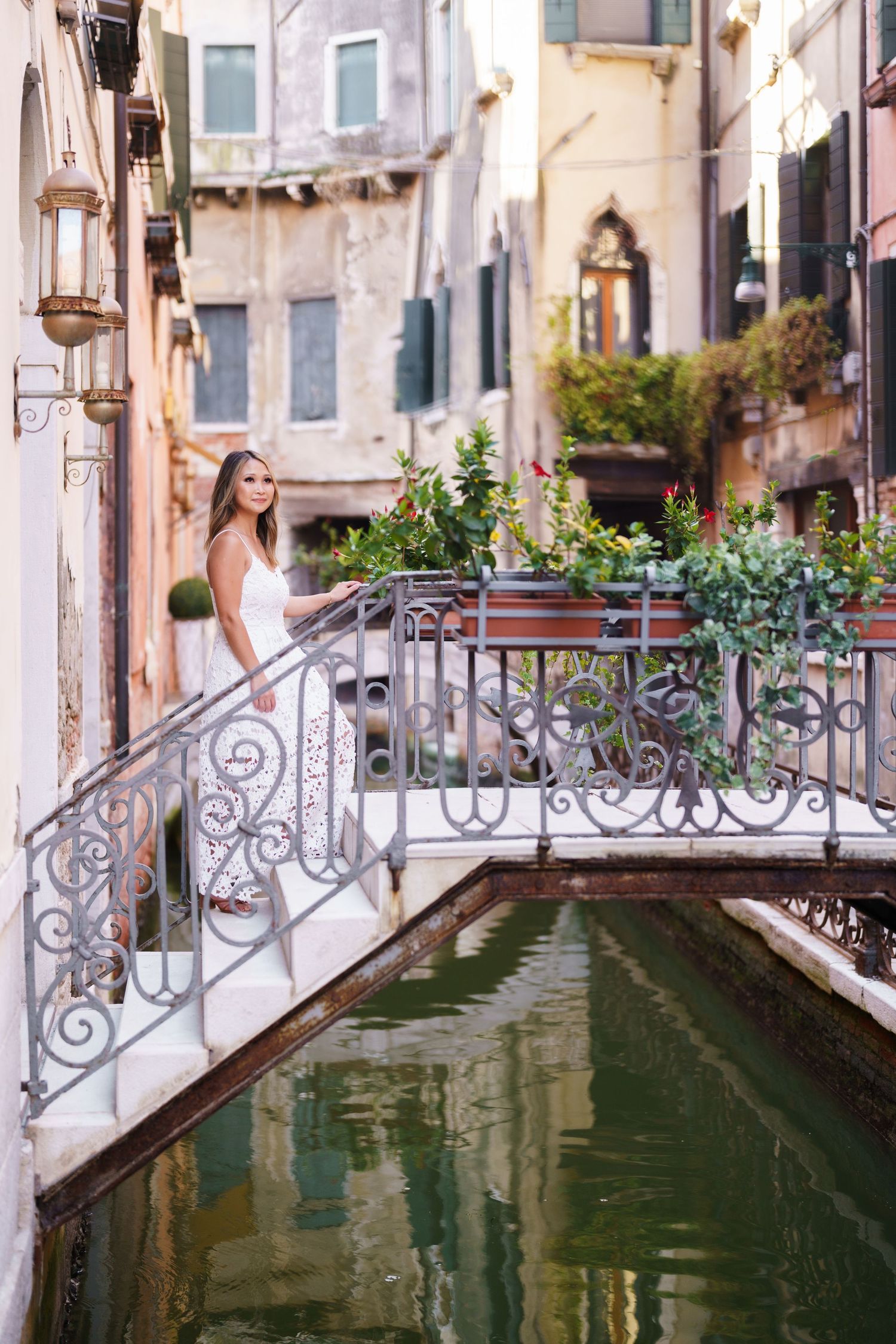 Woman in white dress standing on ornate bridge over green canal in Venice, Italy, surrounded by historic buildings.
