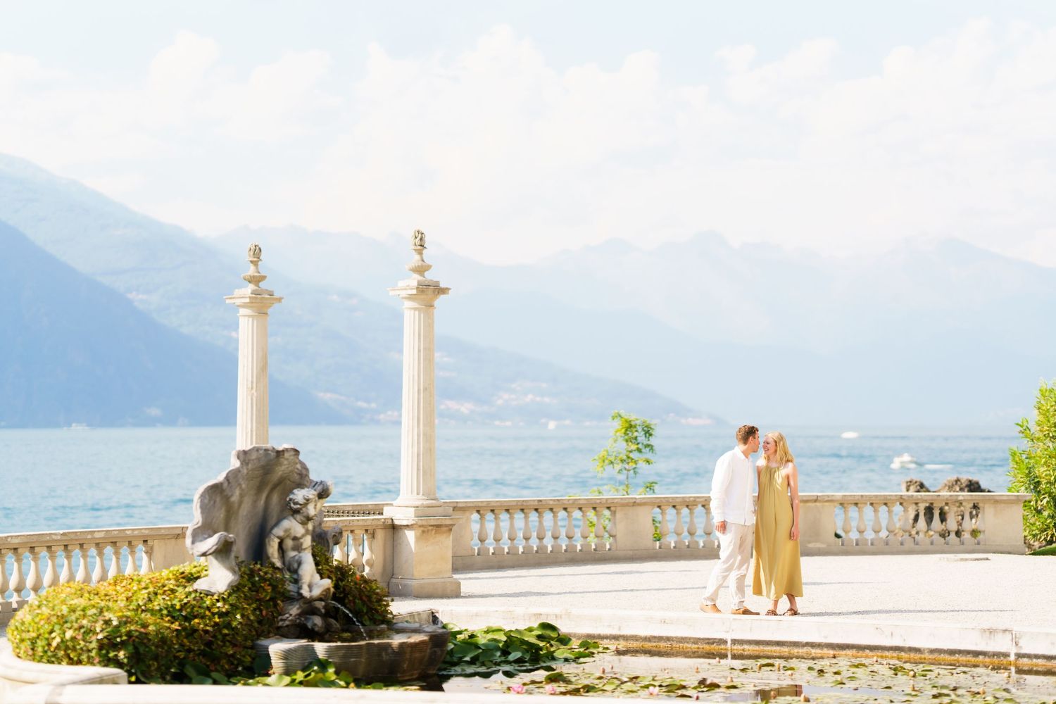 Couple embracing on elegant lakeside terrace with ornate fountain, white balustrade, and mountain backdrop at Lake Como, Italy.