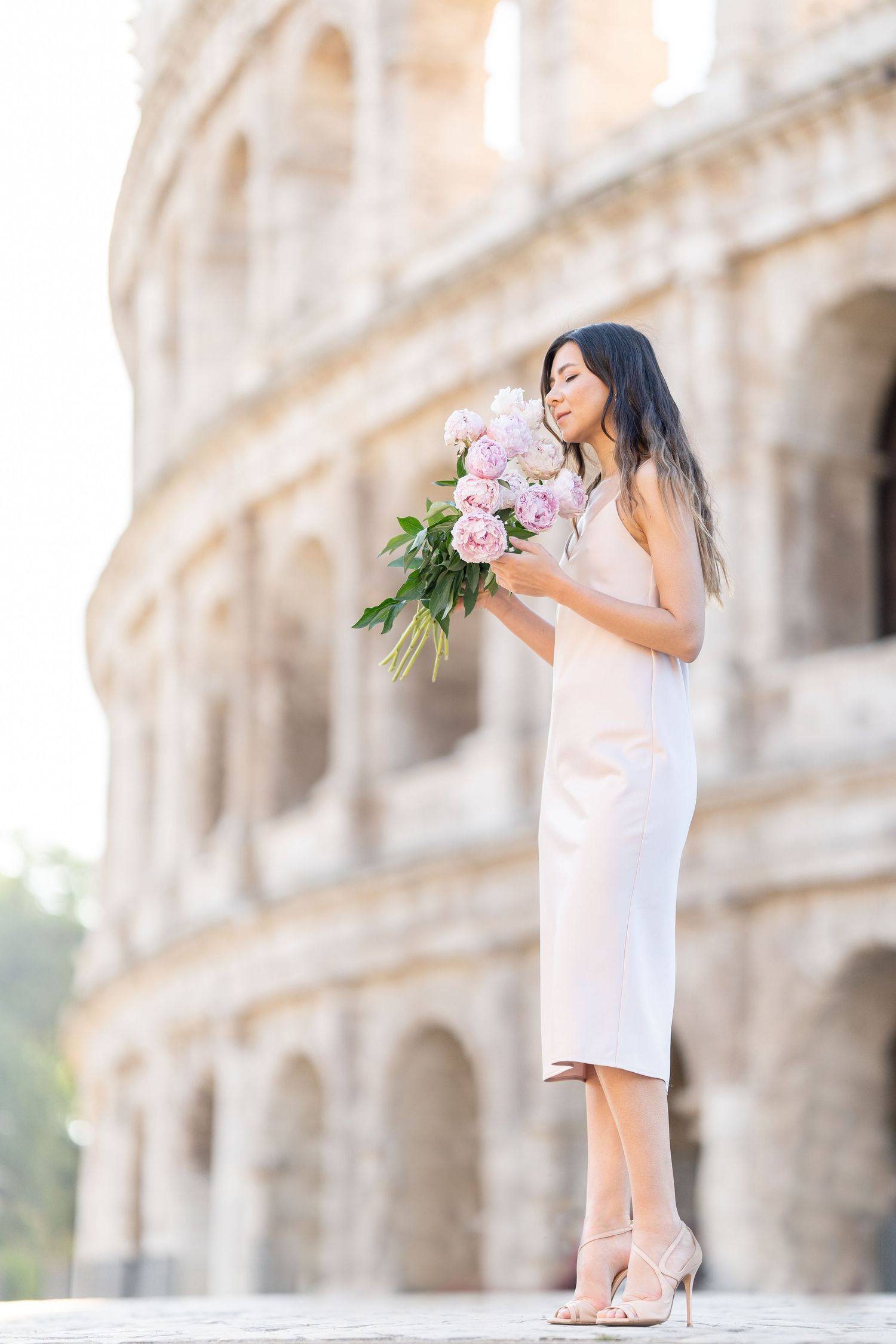 Woman in white dress holding pink flowers bouquet standing in front of the Colosseum in Rome, Italy.