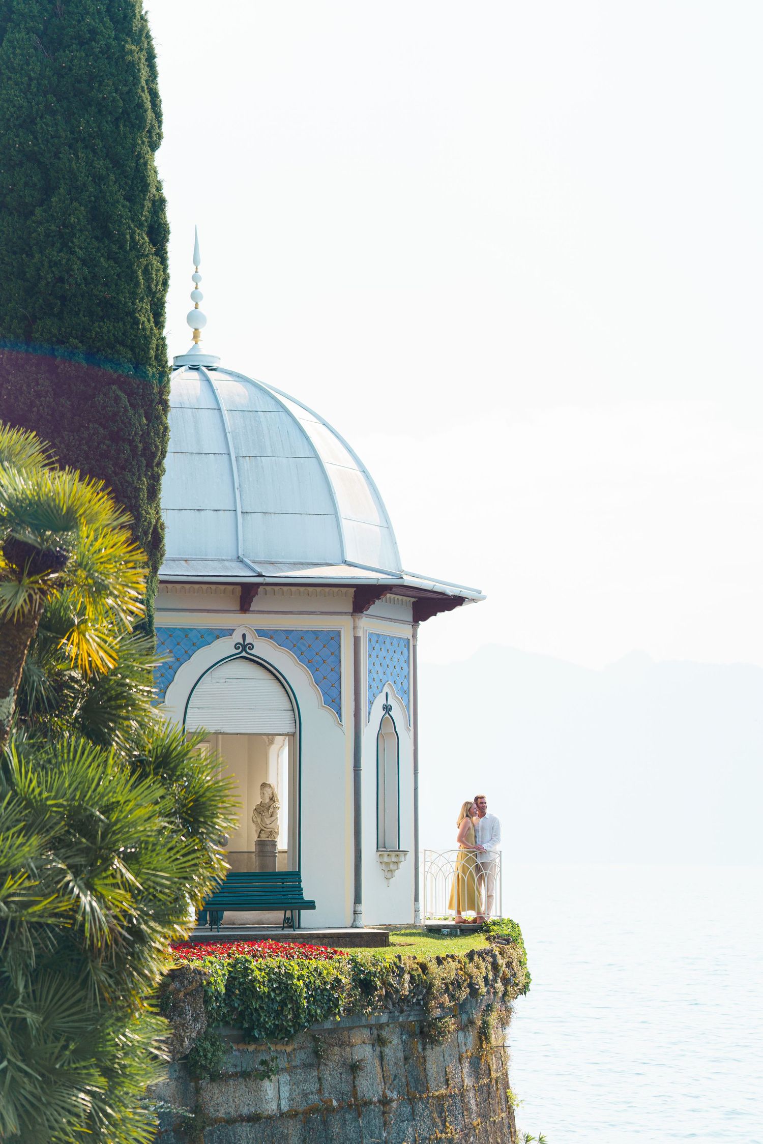Couple embracing in a white domed pavilion overlooking a serene lake, surrounded by lush palm trees and colorful gardens.