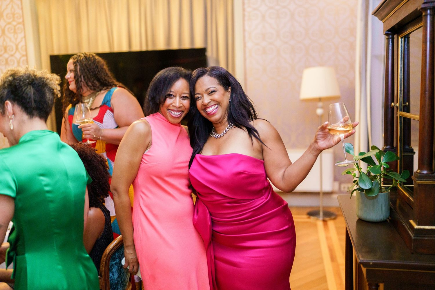 Two women in vibrant pink dresses smiling and holding drinks at an elegant indoor social gathering.