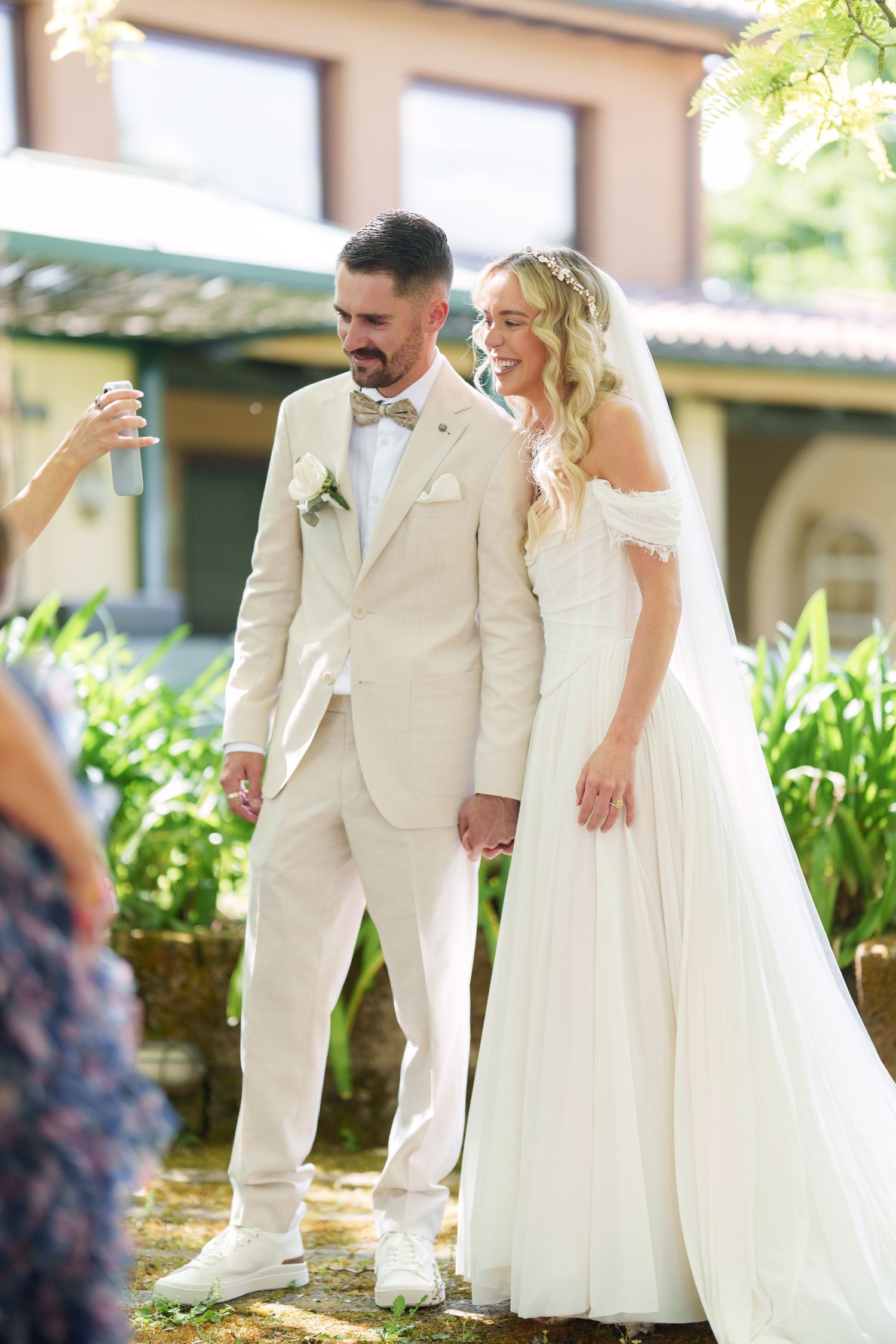 Bride in white off-shoulder gown and groom in cream suit hold hands outdoors at elegant wedding venue.