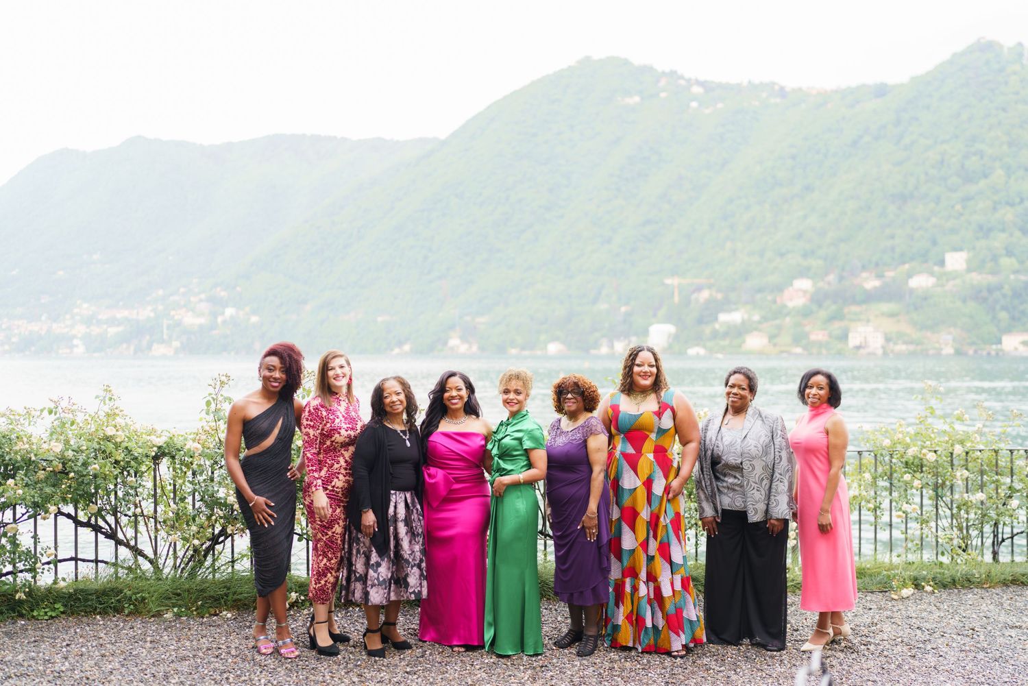 Group of elegantly dressed women posing together outdoors by a scenic lake with mountains in the background.