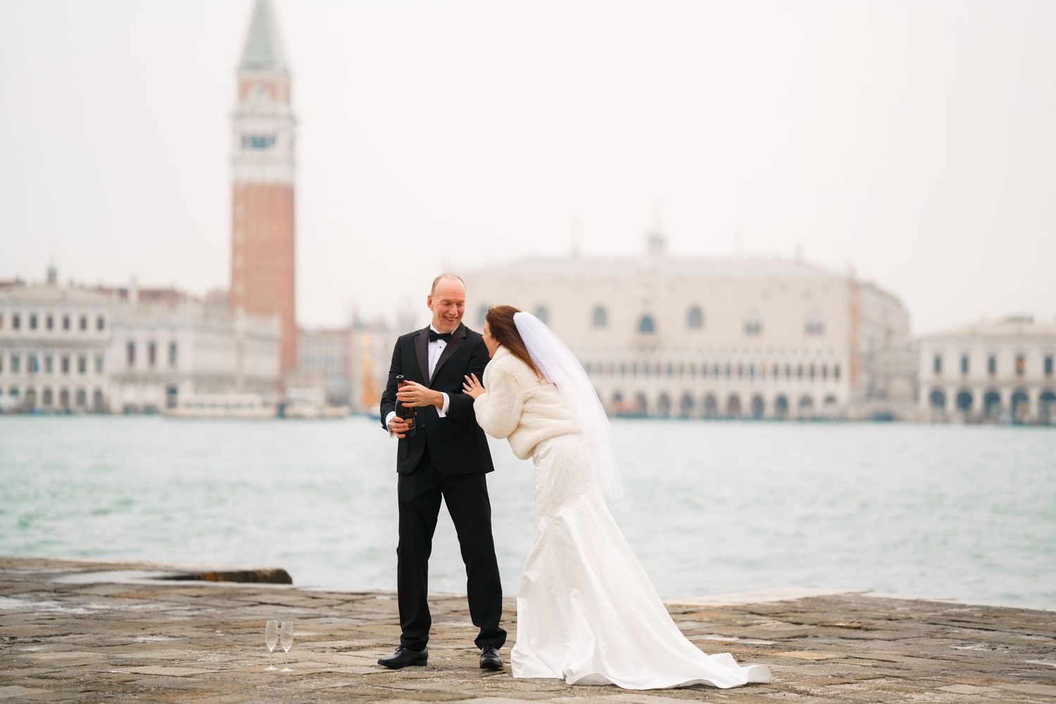 A romantic wedding couple poses on a stone bridge overlooking Venice's iconic waterfront and historic buildings.