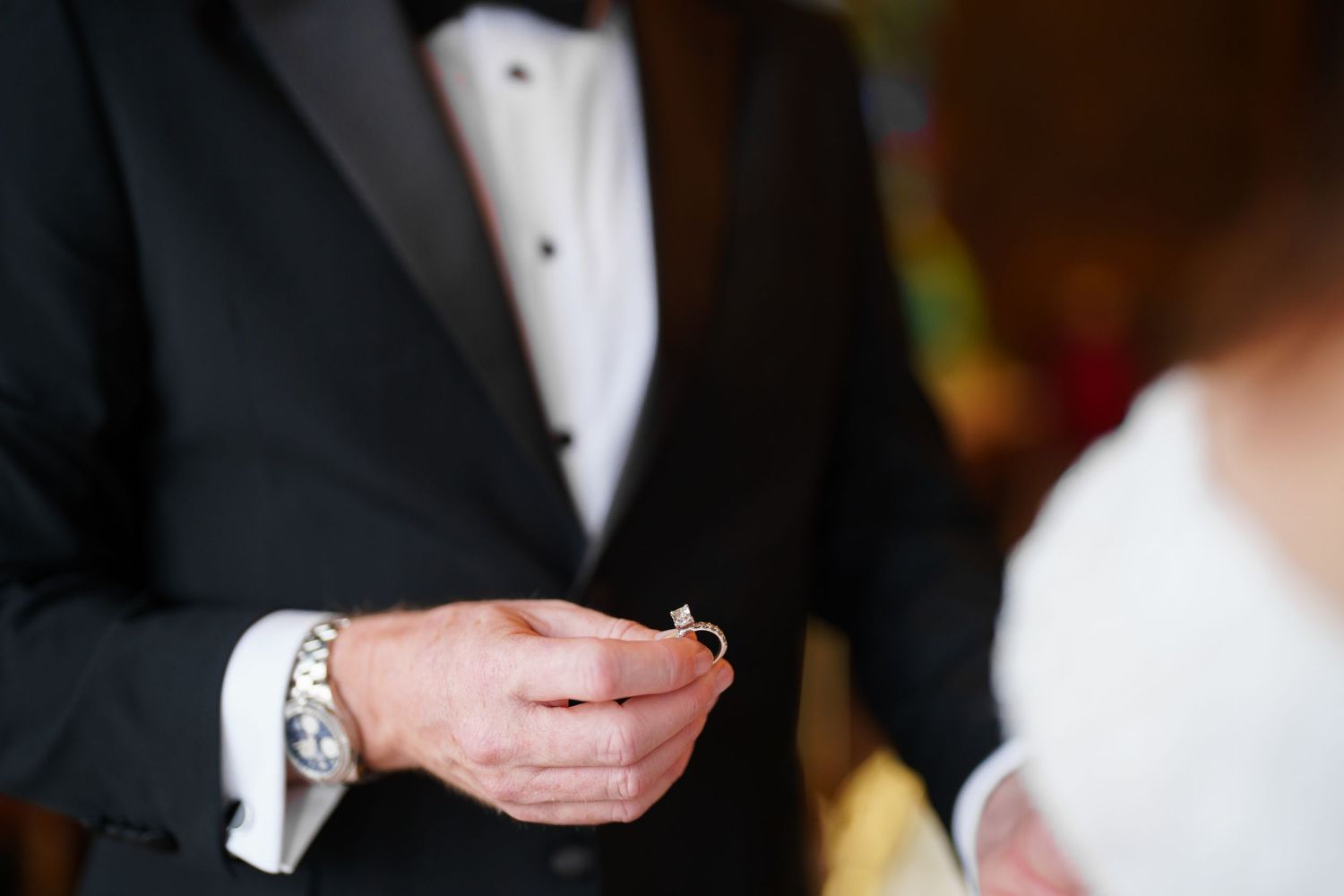Close-up of groom in black tuxedo holding wedding ring during ceremony.
