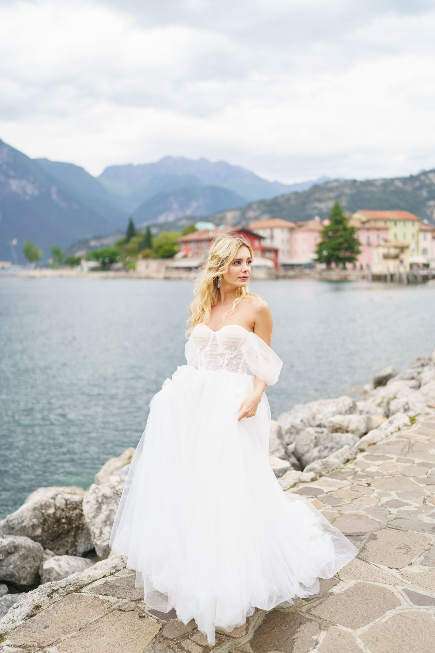 A white wedding dress flows elegantly against a lakeside mountain backdrop in a scenic European village setting.