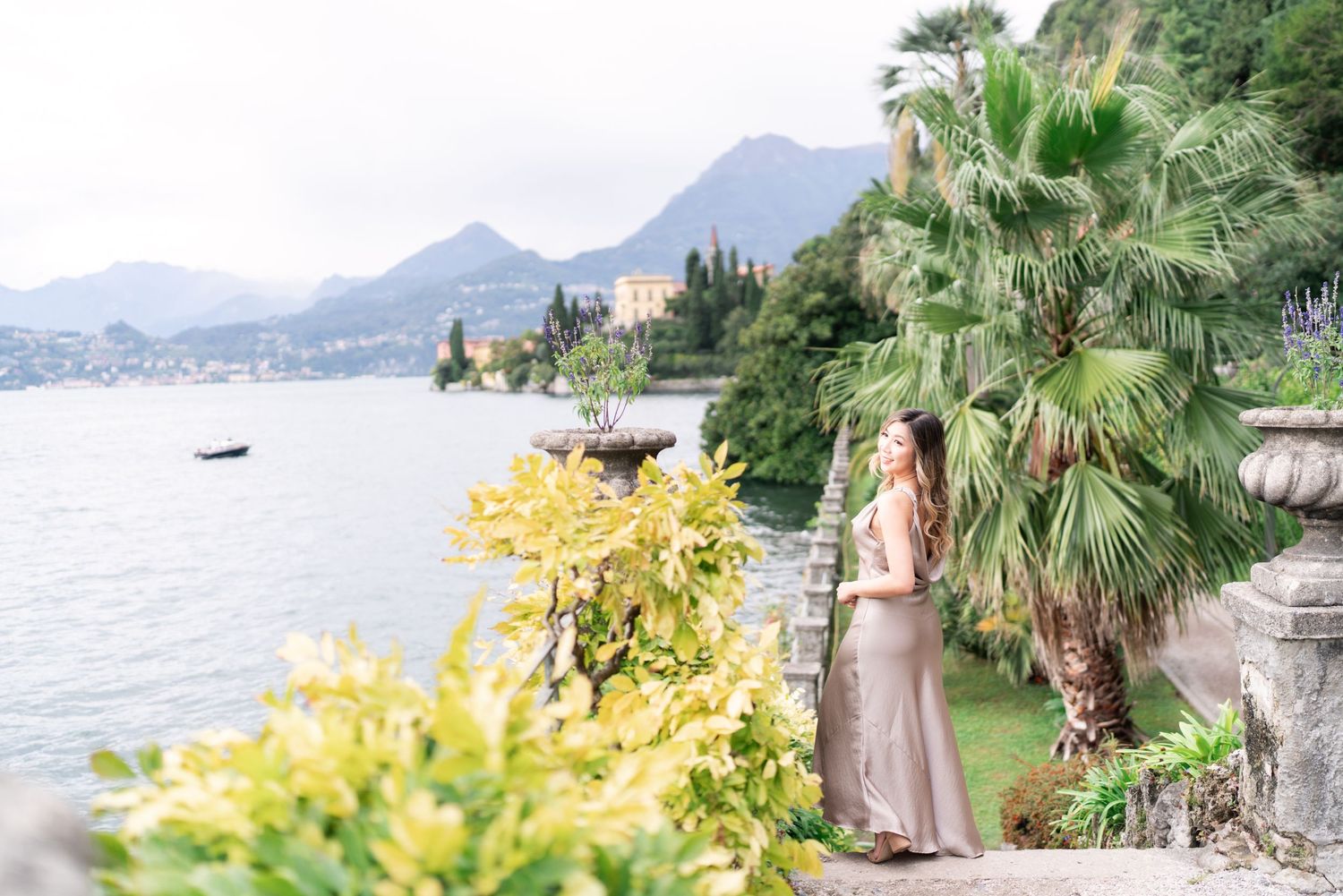 A scenic coastal view of Lake Como in Italy with mountains, palm trees and yellow flowering bushes along the shoreline.