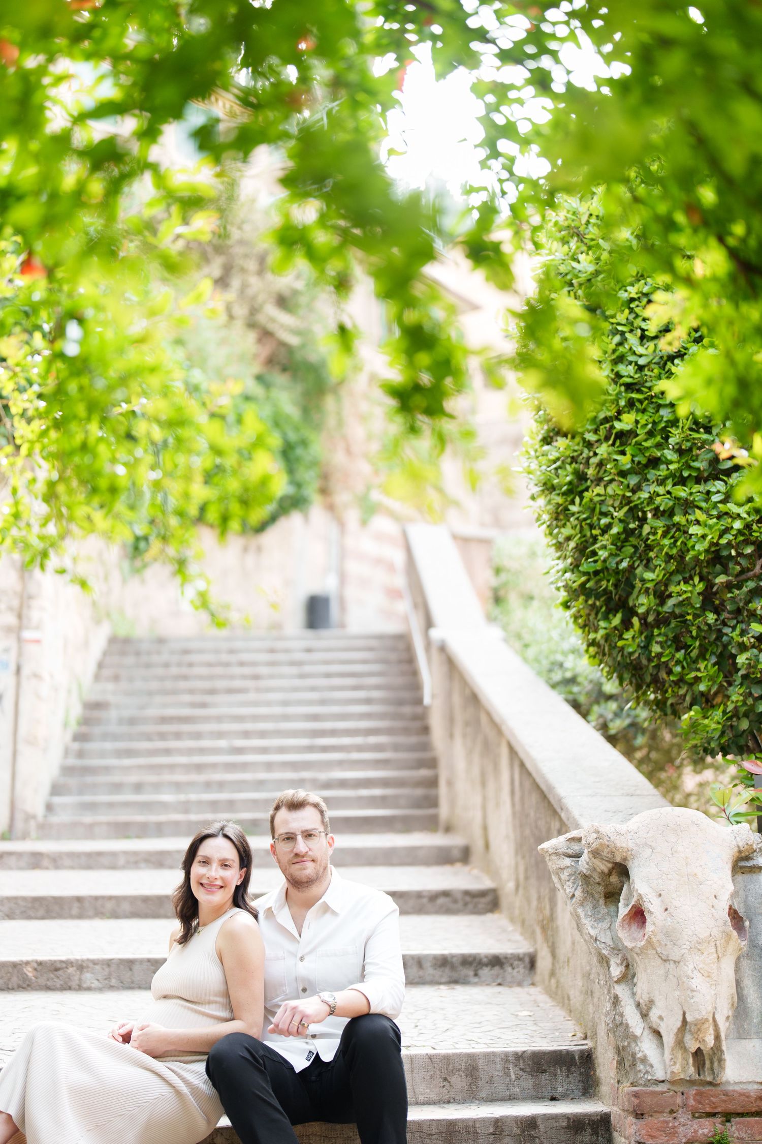 A couple sits together on stone steps surrounded by lush green trees and foliage in a romantic outdoor setting.