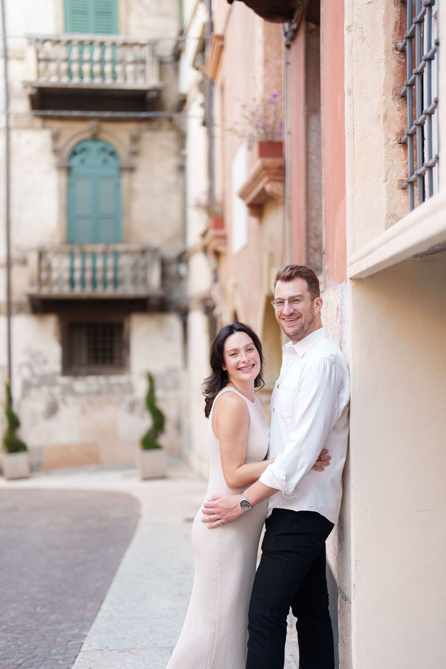 Romantic couple embraces and smiles while standing against a rustic stone wall in an old European street setting.