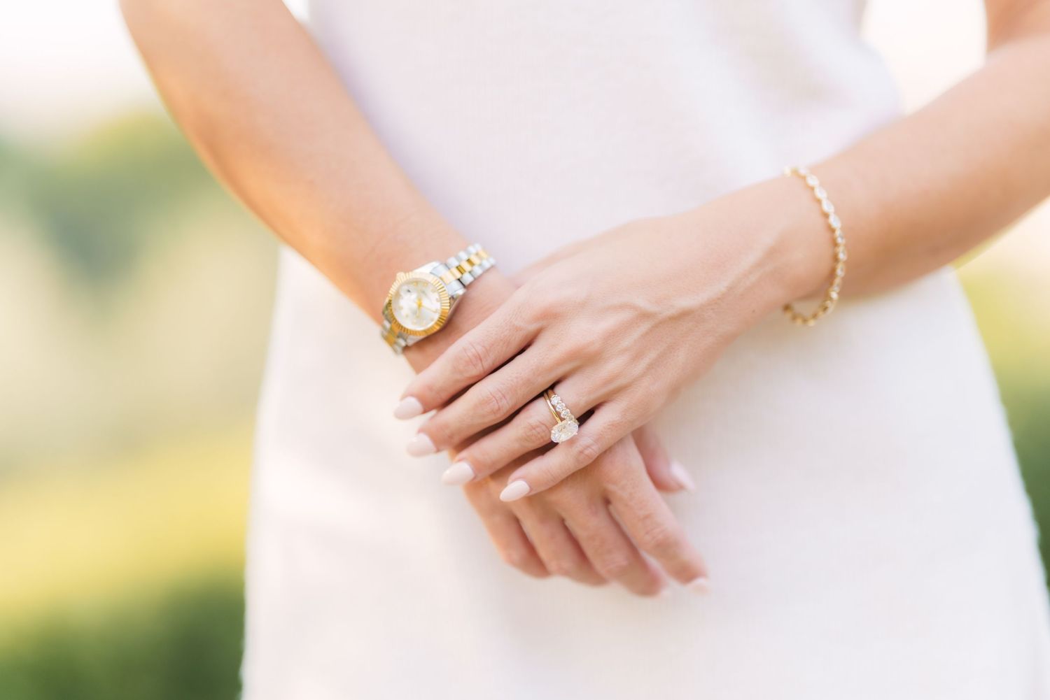 Close-up of hands wearing an elegant gold watch, bracelet and engagement ring with a white dress in soft natural light.