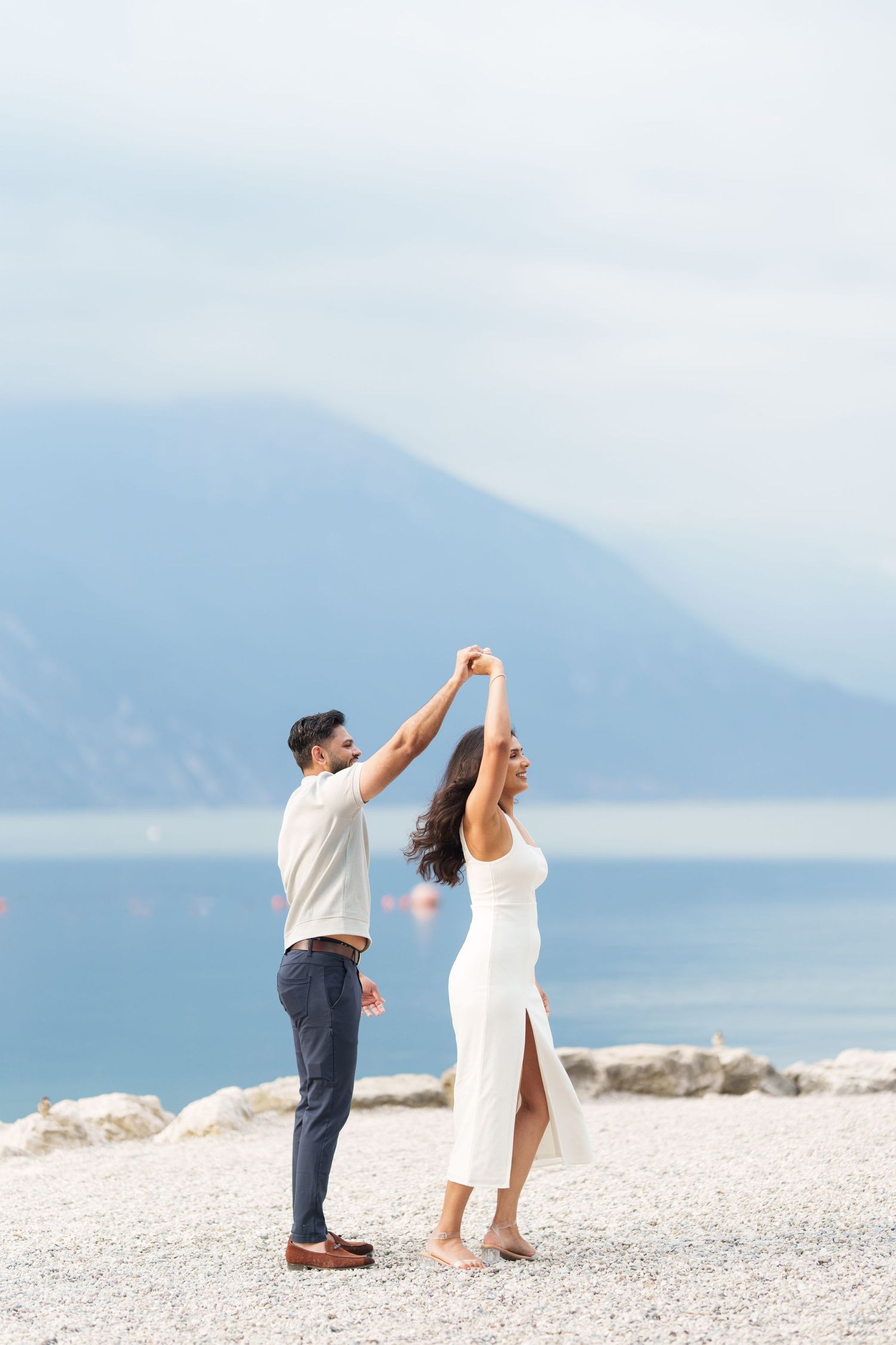 Two people in white and dark outfits dance joyfully on a scenic pebble beach with mountains and water in the background.