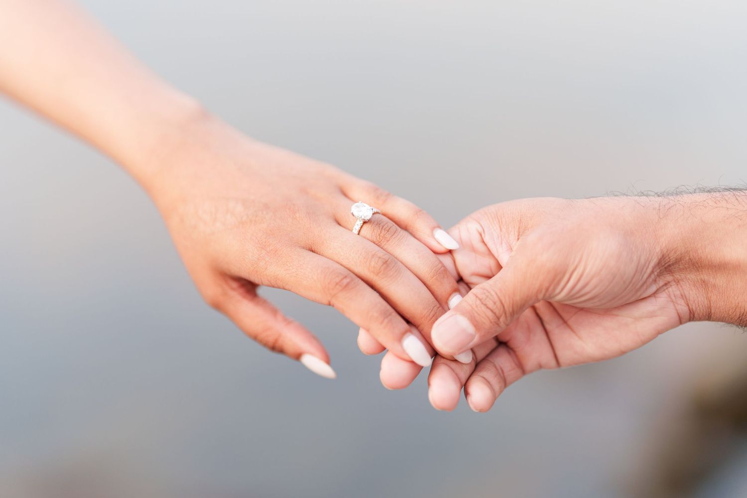 Two hands gently touch with one wearing an elegant engagement ring against a soft blurred background.