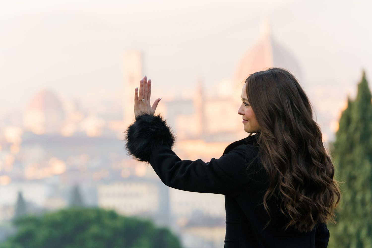 A person with long dark hair stands with raised arms against a blurred city skyline at sunset.