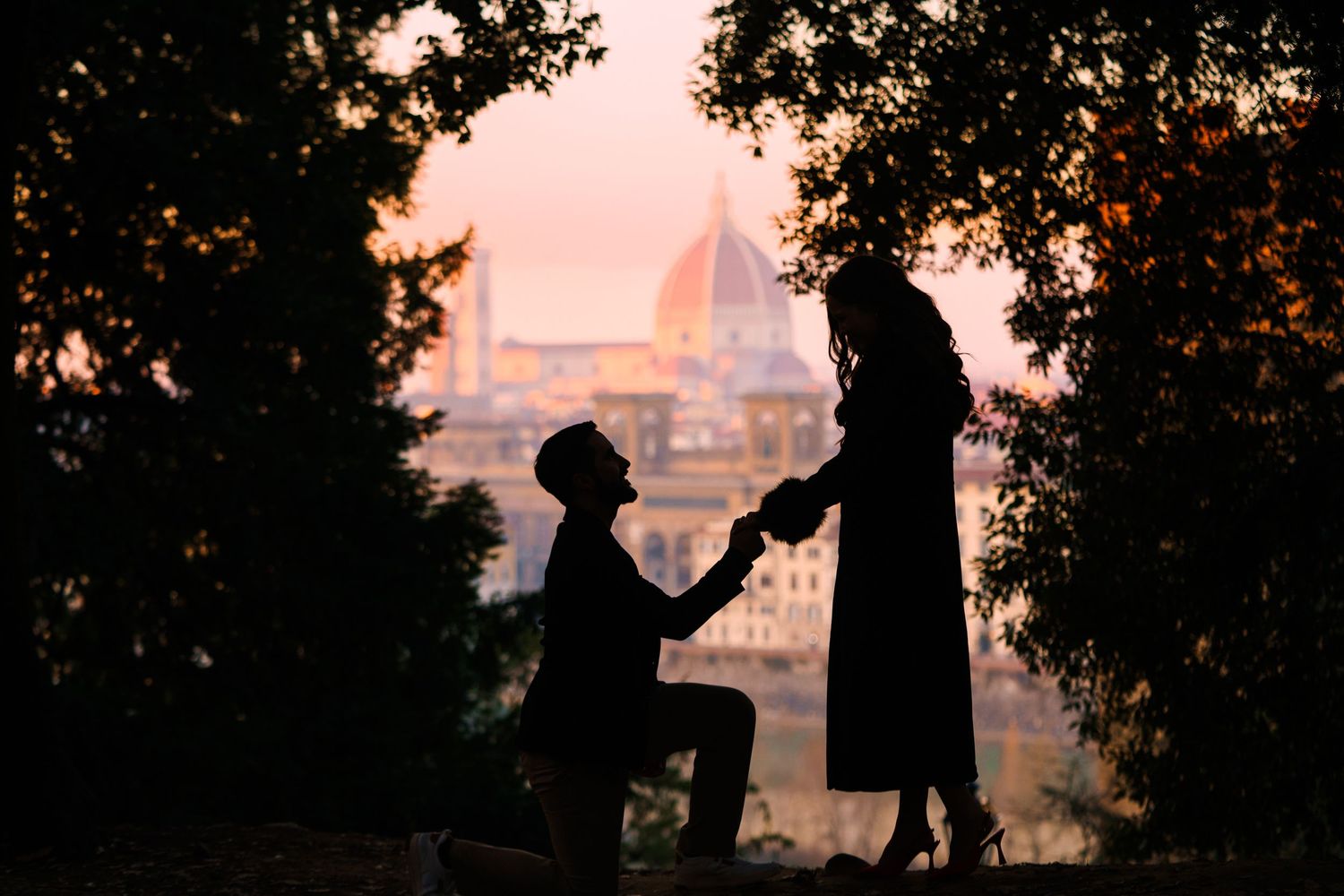 Silhouettes of people standing on a hilltop against a pink sunset sky with city buildings visible in the background.