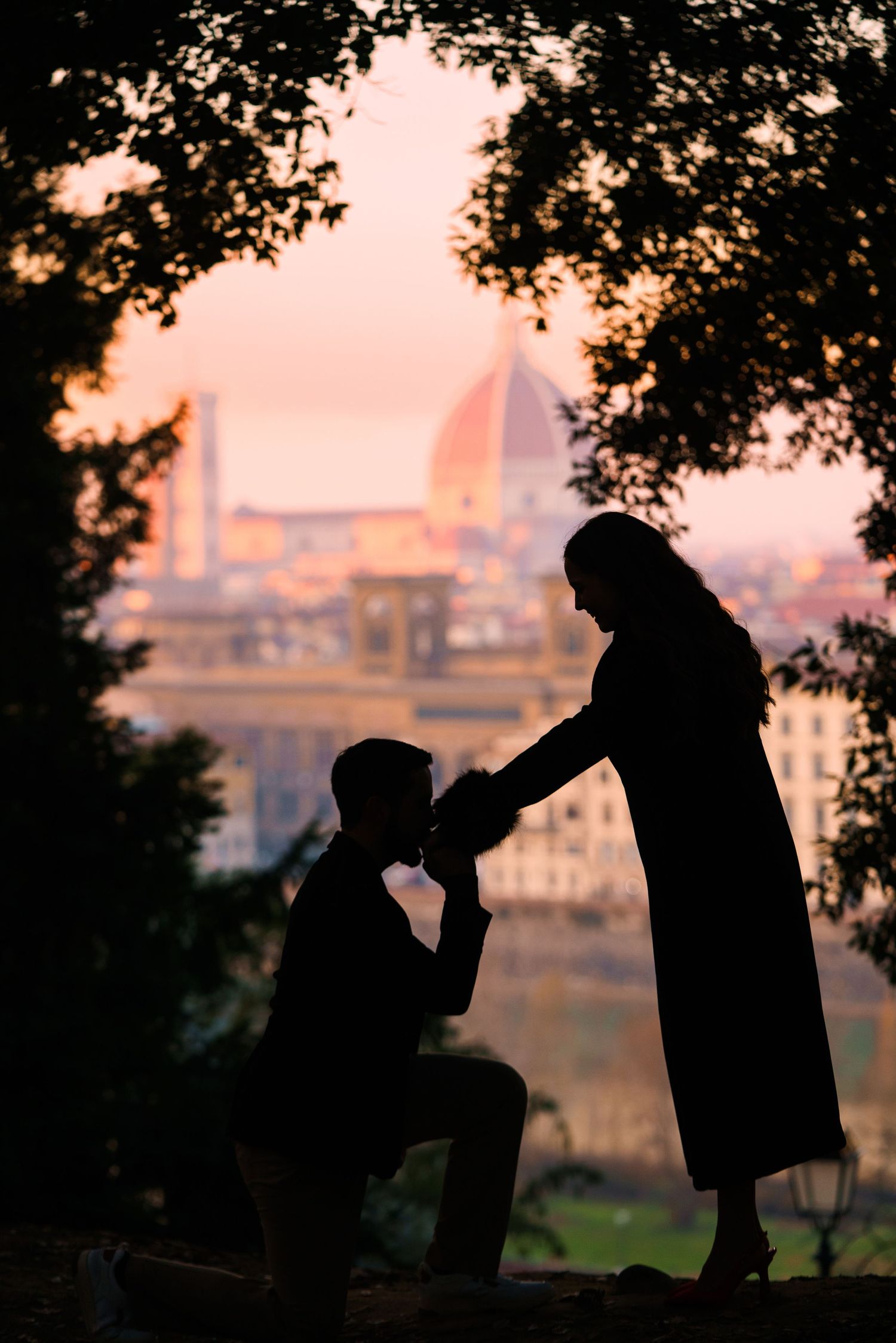 Two silhouettes share a romantic moment against a sunset cityscape backdrop, creating a dreamy atmospheric scene.